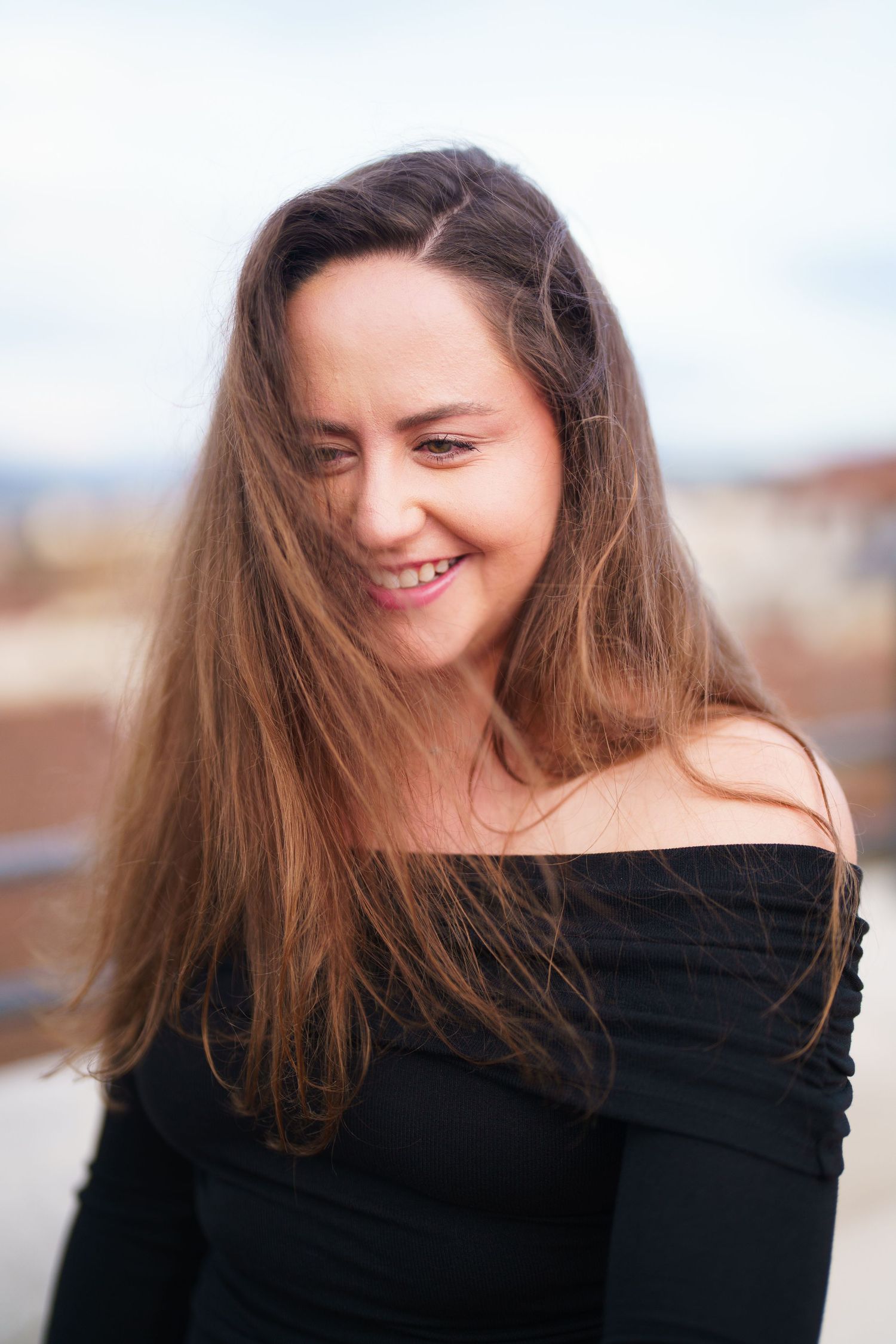 Portrait of a smiling person with long brown hair wearing a black off-shoulder top against a blurred background.
