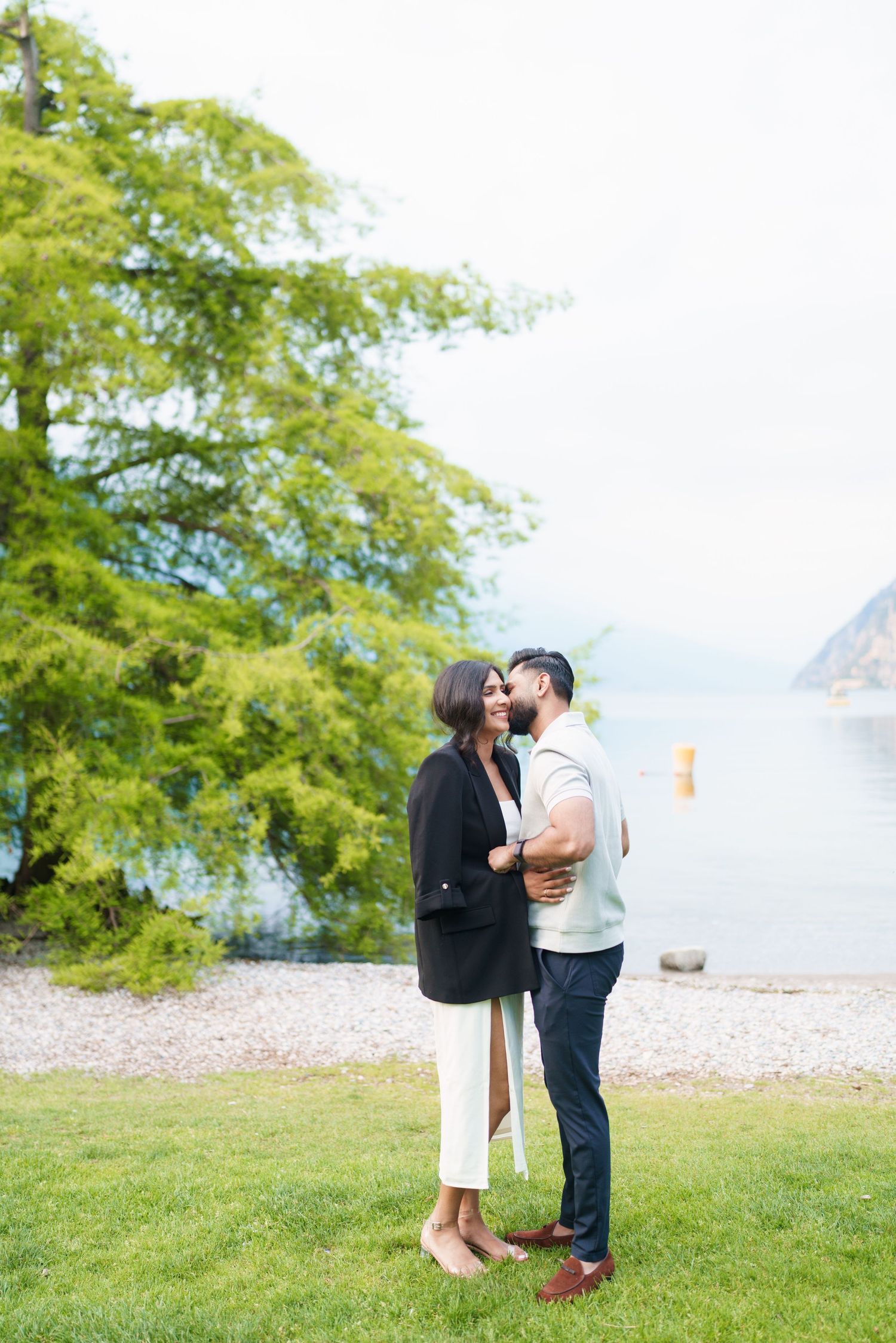 A couple shares romantic moments by a scenic lake with mountains and green trees in the background.