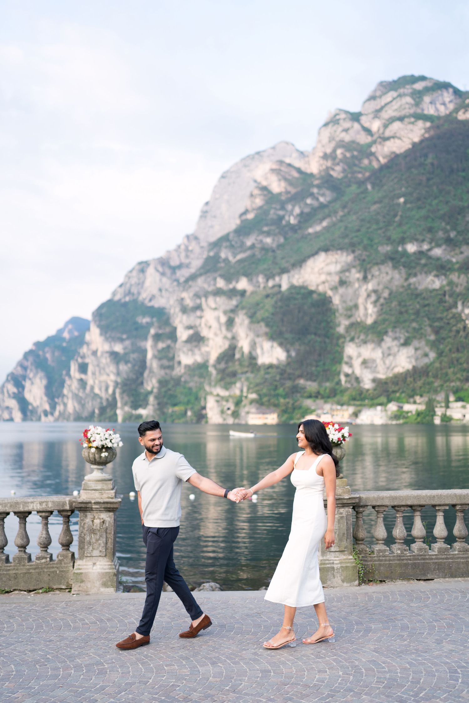A couple dances playfully along a lakeside promenade with scenic mountain cliffs in the background at Lake Garda, Italy.