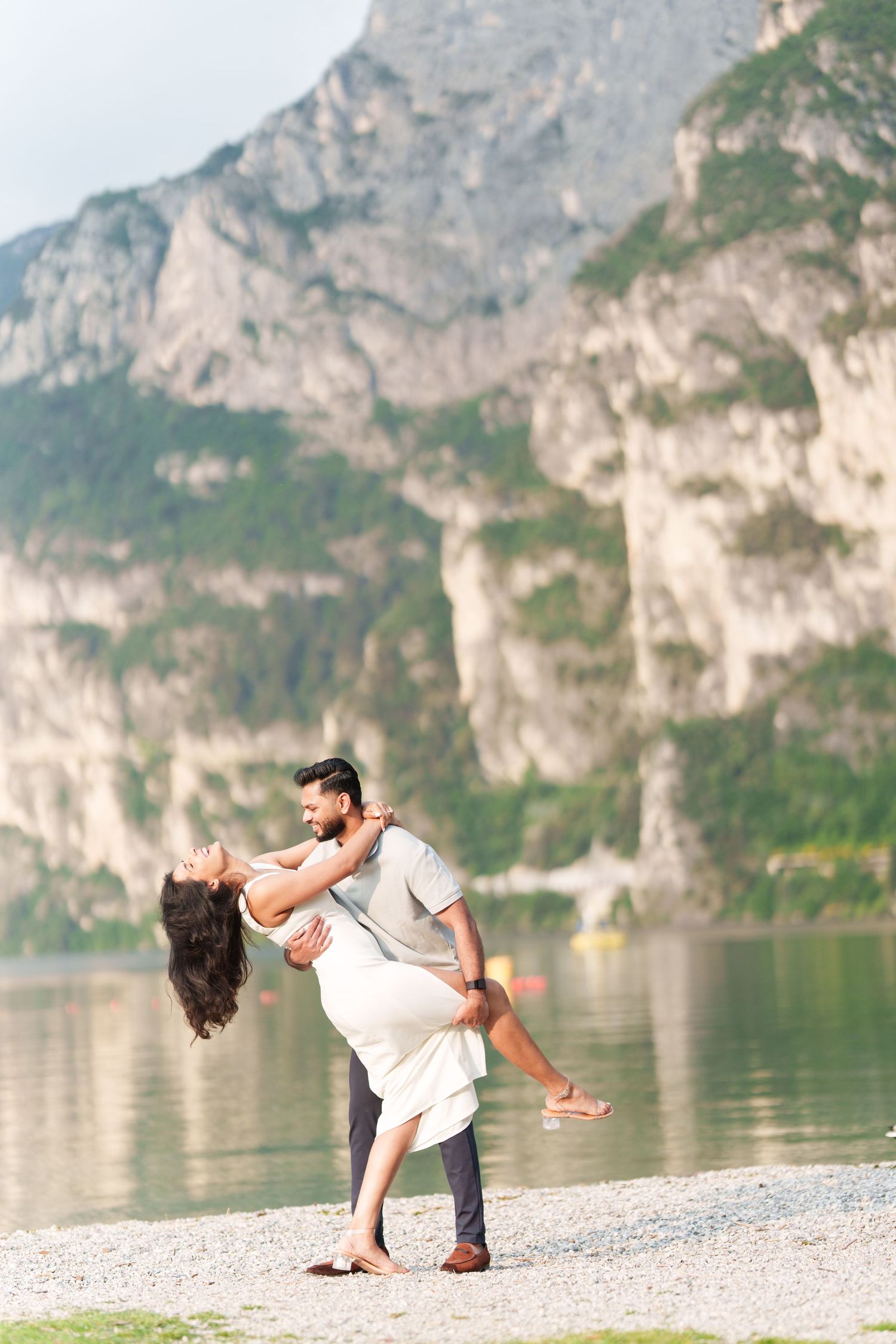 A couple performs a romantic dance pose on a scenic lakeside beach with dramatic mountain cliffs in the background.