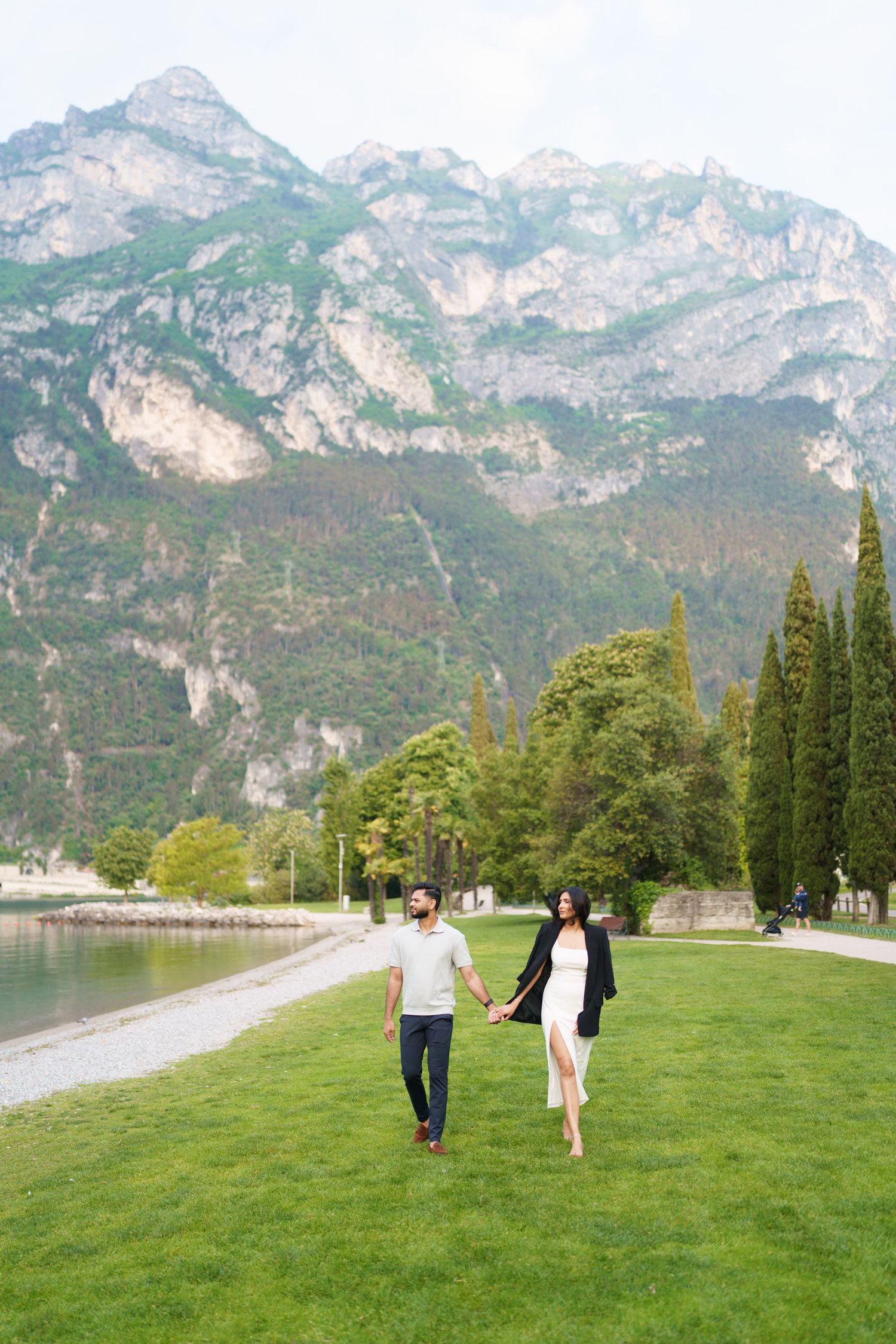Two people walk hand in hand across a green lawn with dramatic mountain peaks rising in the background near a lake.