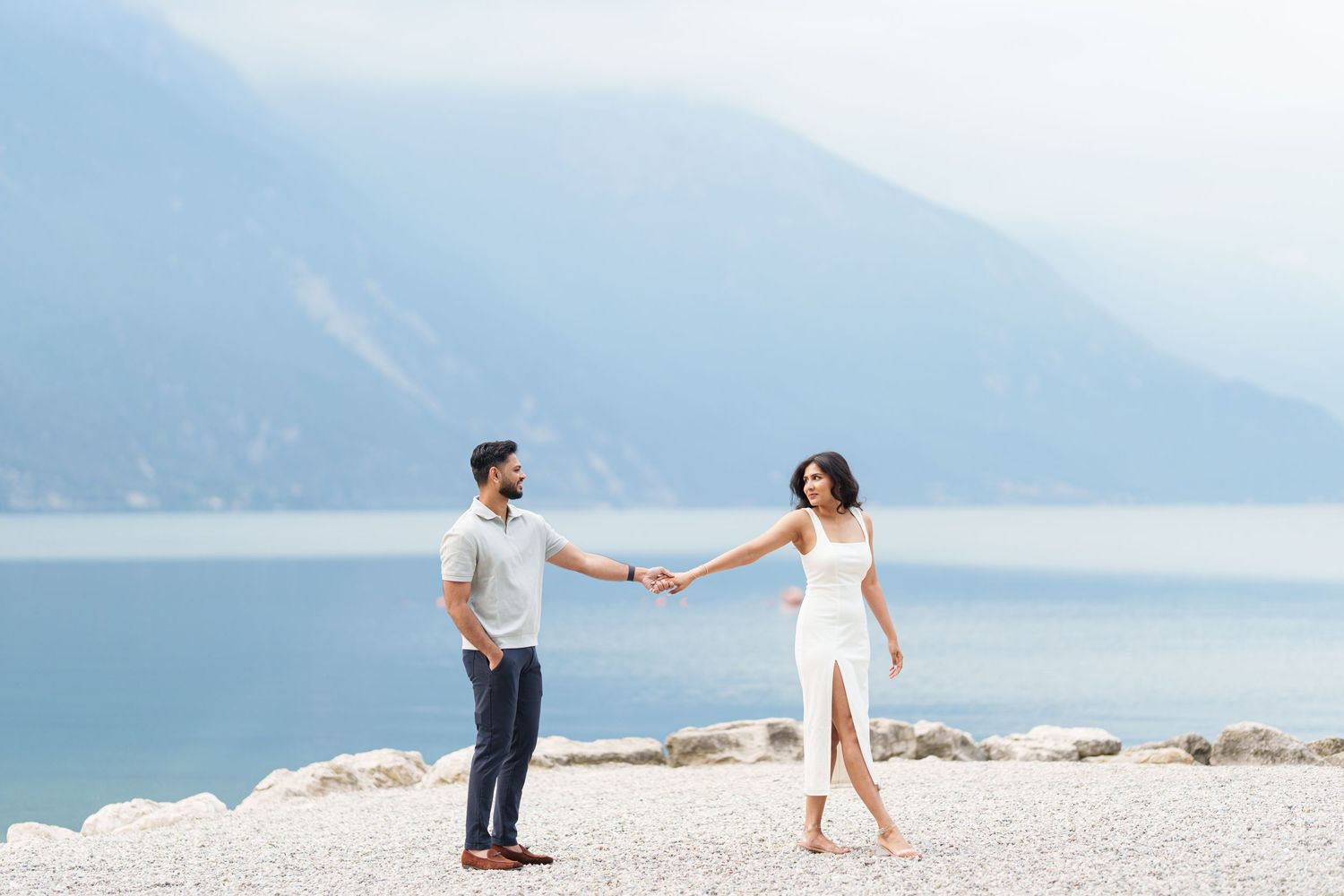 Couple holding hands while walking along a scenic mountain lake shoreline in a romantic outdoor setting.