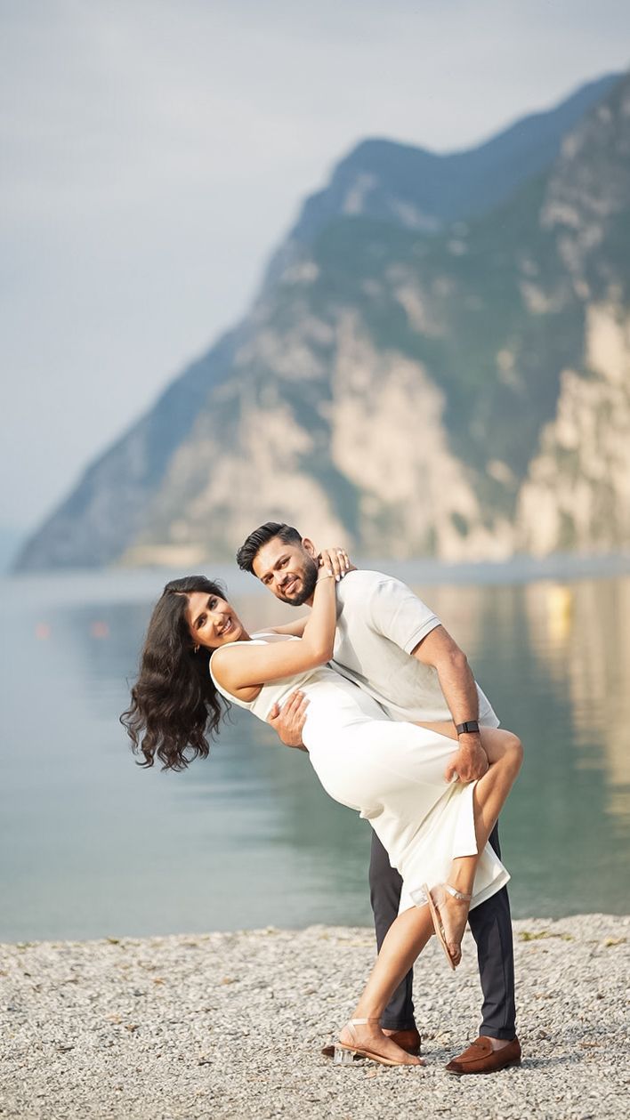 A romantic couple dances playfully on a rocky beach with scenic mountain cliffs and lake in the background.