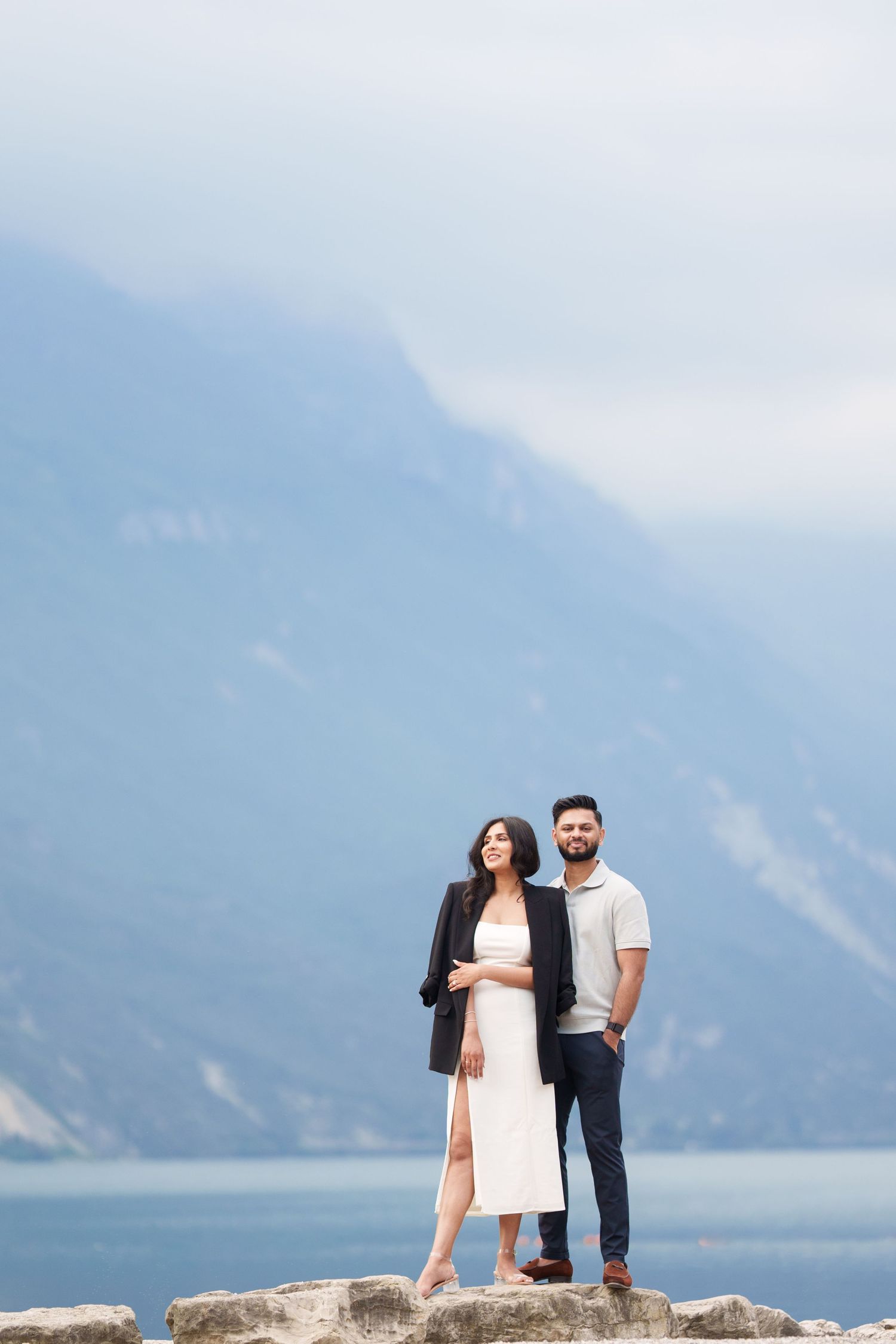 Couple in casual attire standing on rocky coastline with misty mountains and lake in the background.