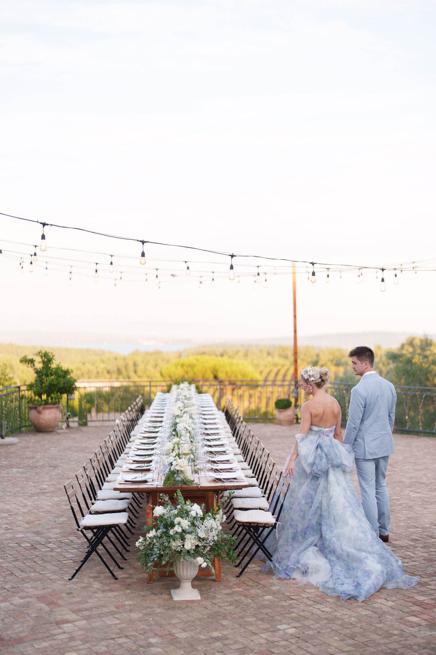 Couple in blue wedding attire standing beside a long outdoor reception table with string lights and vineyard views.