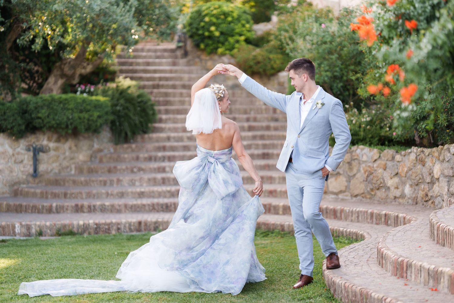 Bride and groom dancing outdoors in a garden, she wears a blue wedding gown, he wears a light gray suit.