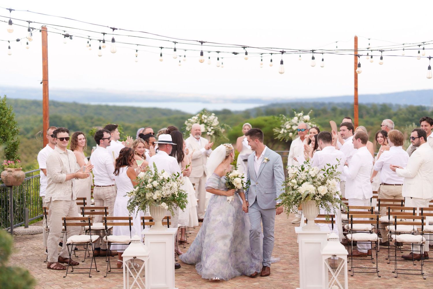 Outdoor wedding ceremony on a scenic overlook with string lights, white floral arrangements, and guests dressed in white attire.