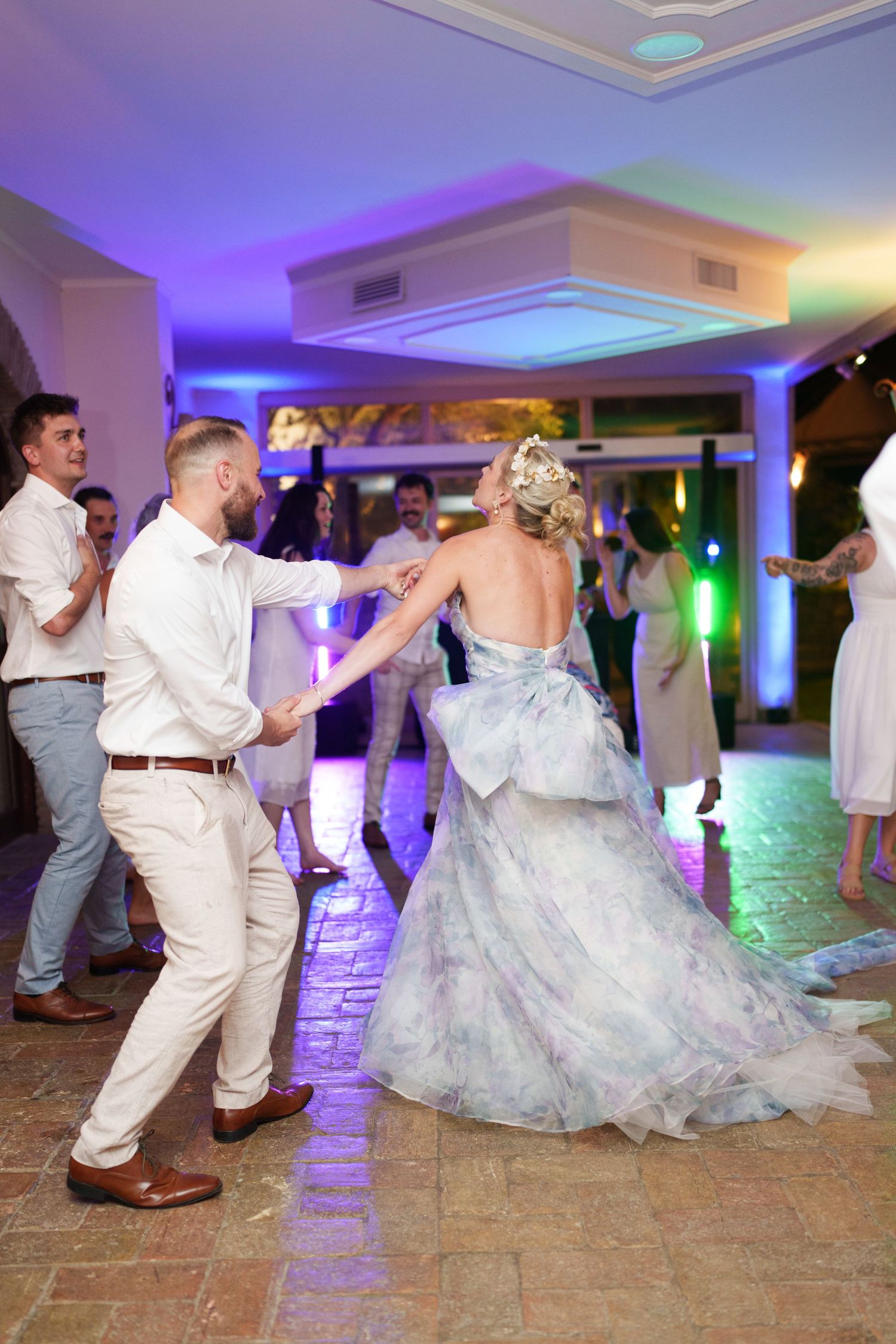Bride and groom dancing together at wedding reception with guests celebrating in colorful venue lighting.