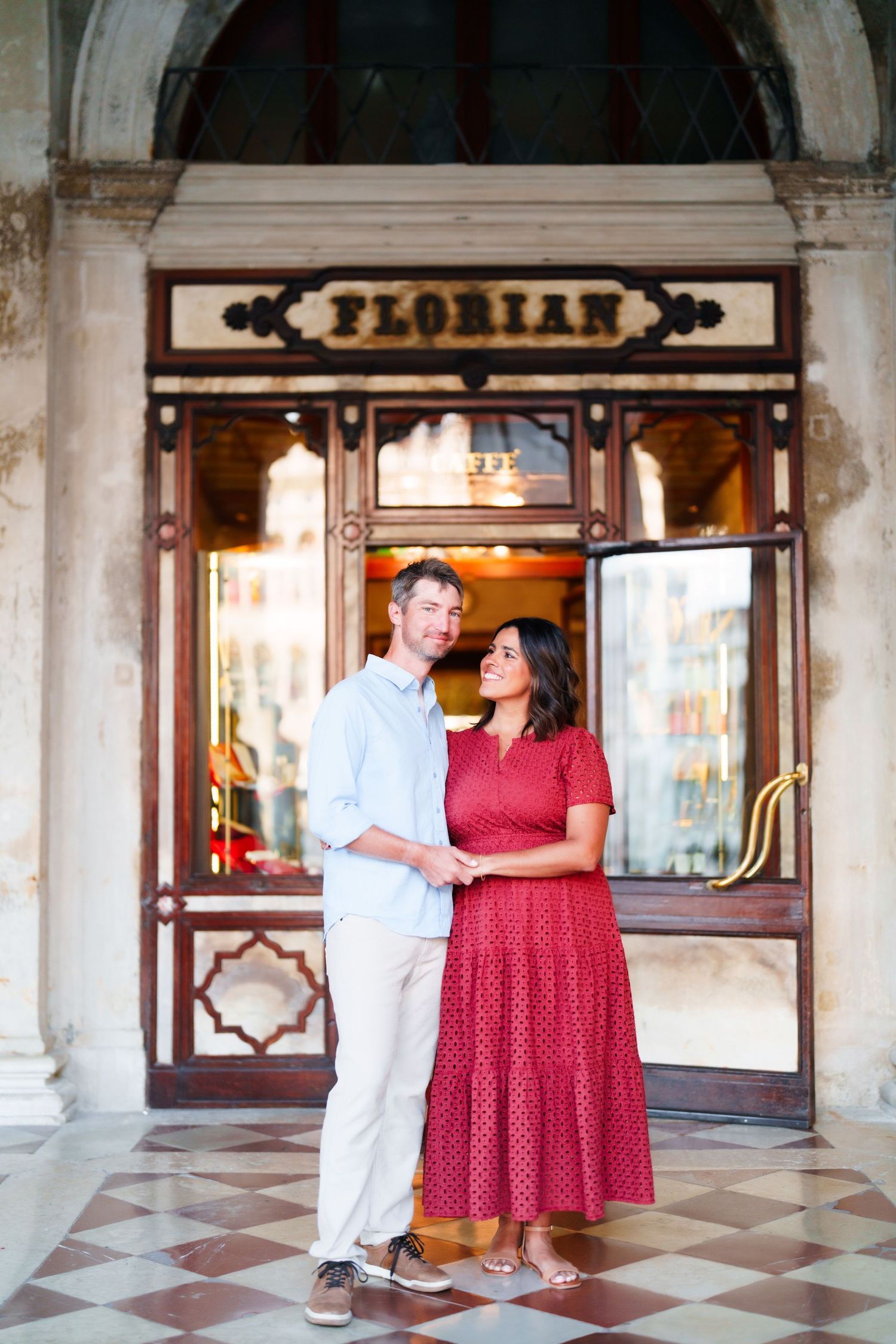 Couple posing and smiling in front of Caffe Florian in Venice, woman in red dress, man in light blue shirt.