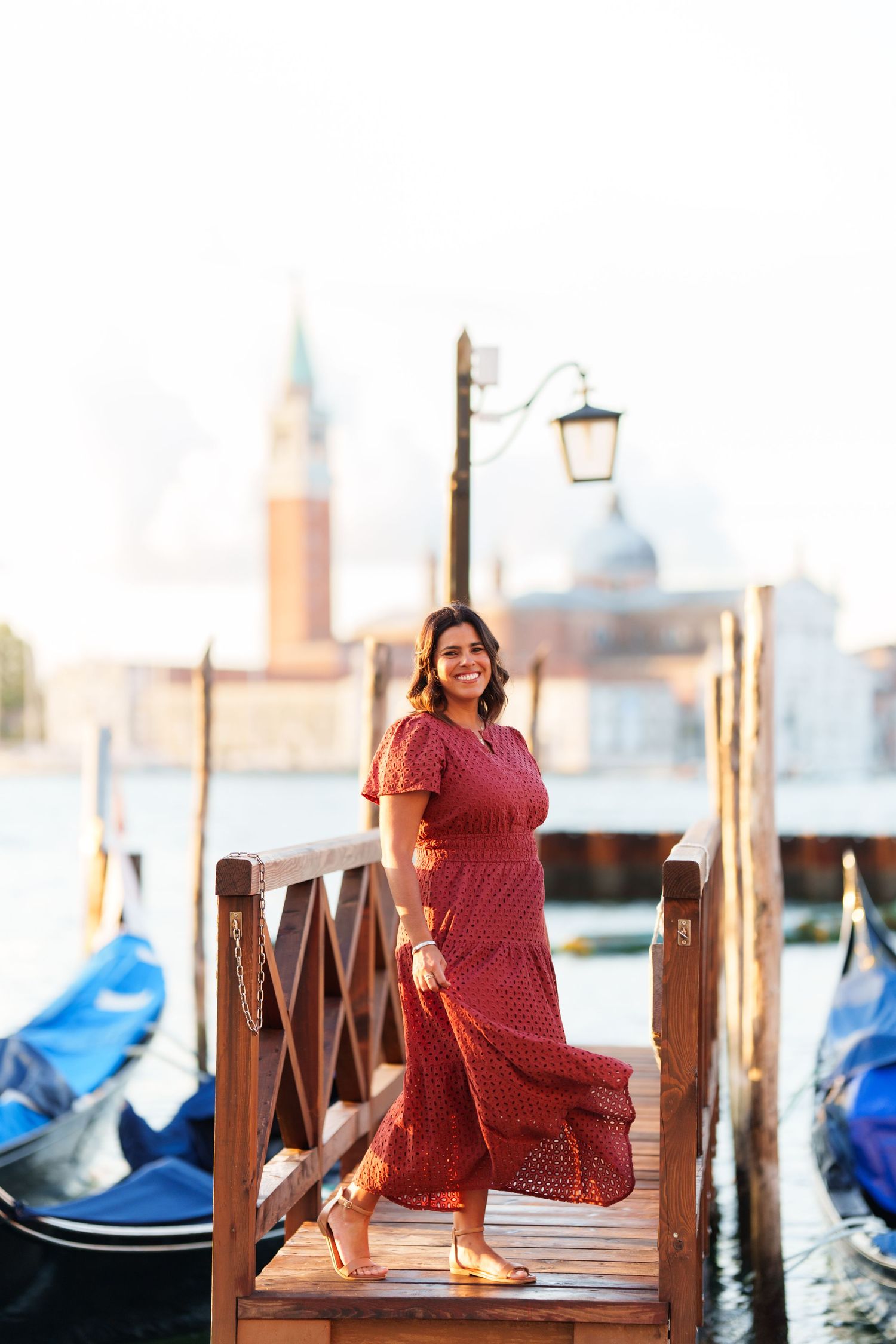 Woman in red dress smiling on Venice gondola dock with San Giorgio Maggiore church in background.