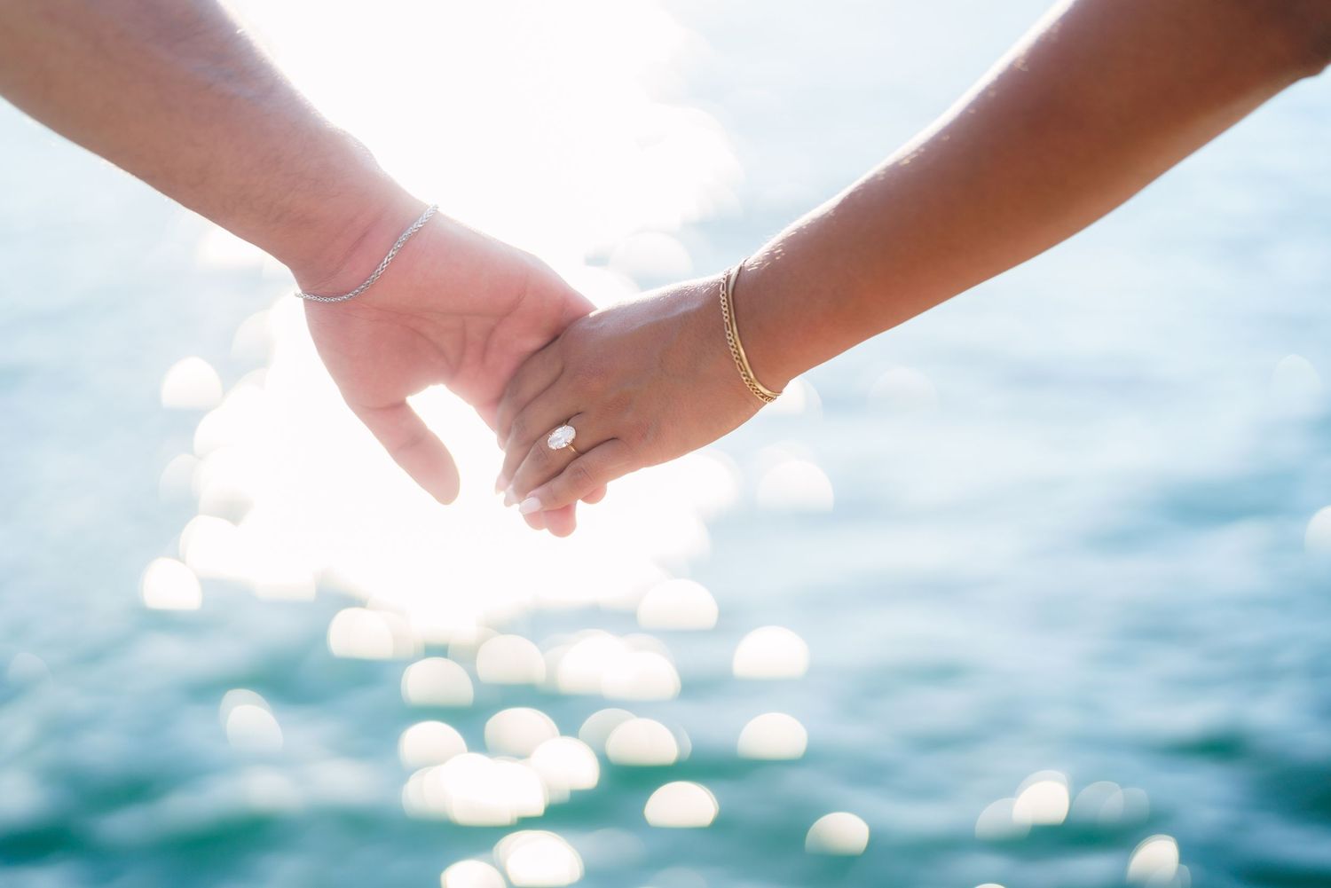 Couple holding hands with sparkling ocean bokeh background, woman wearing gold bracelet and engagement ring.