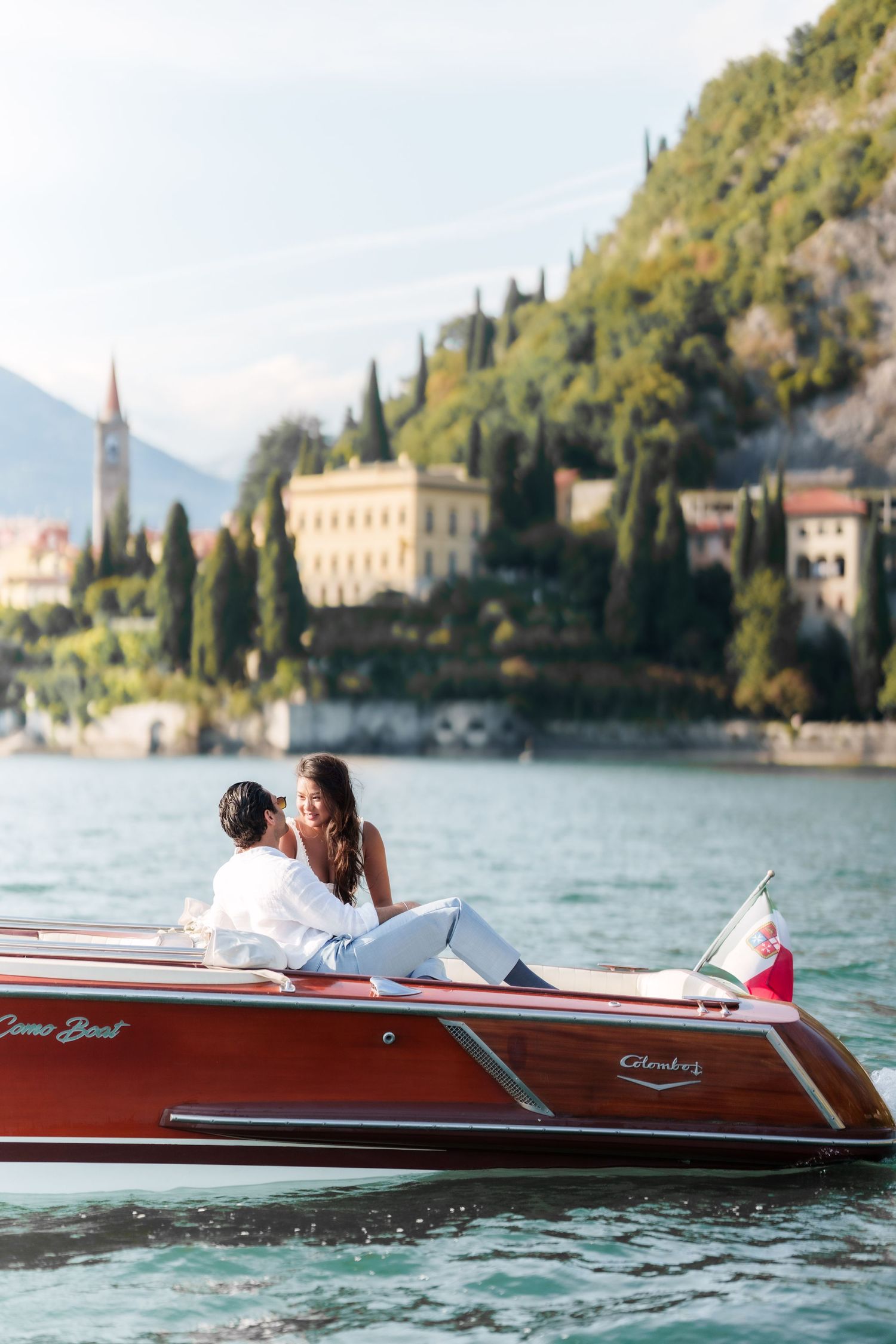 Couple relaxing on a classic wooden Como Boat on Lake Como, Italy, with lush hills and elegant villas in the background.