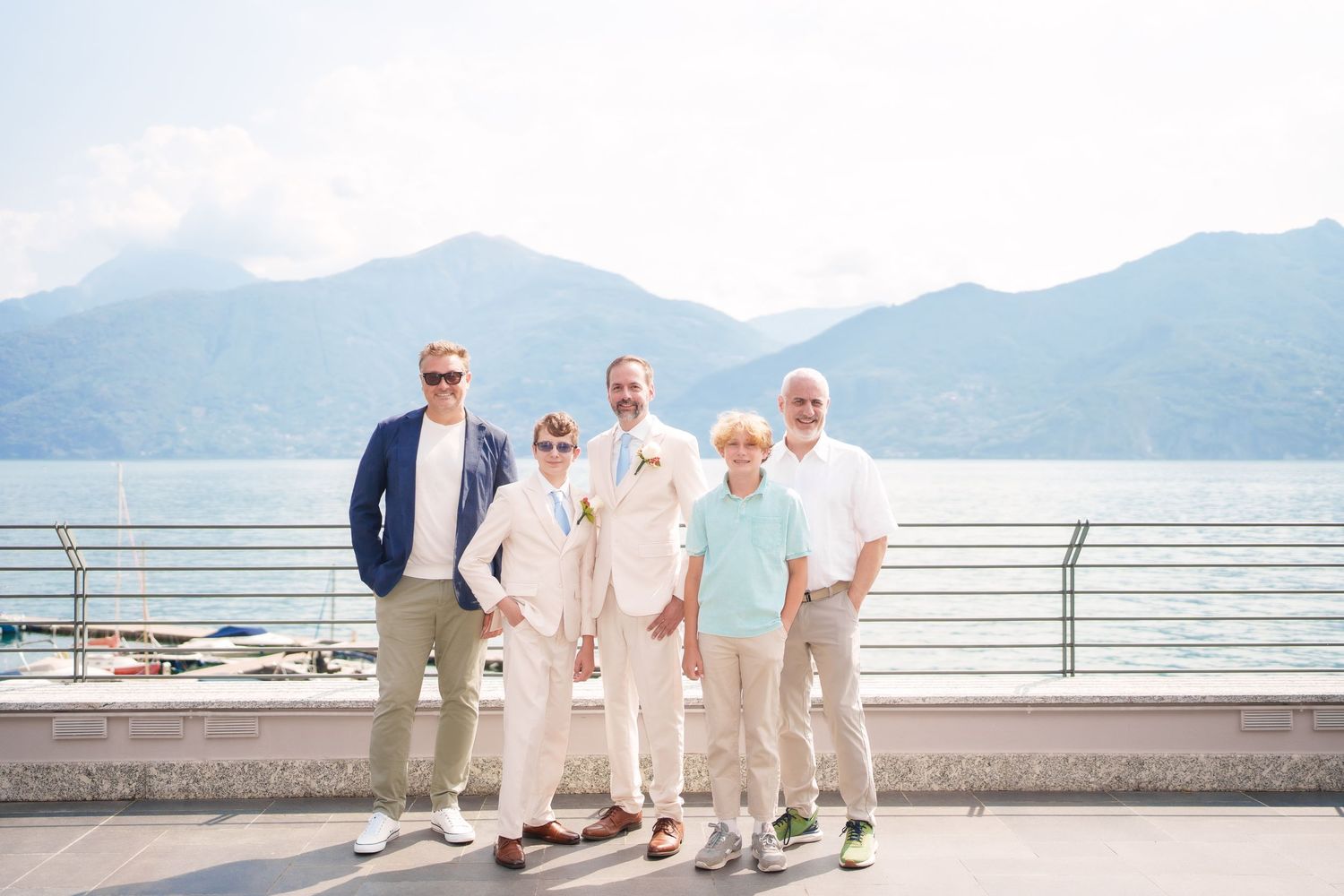 Five men posing together on a lakeside terrace with mountain backdrop, dressed in elegant summer attire for a wedding celebration.