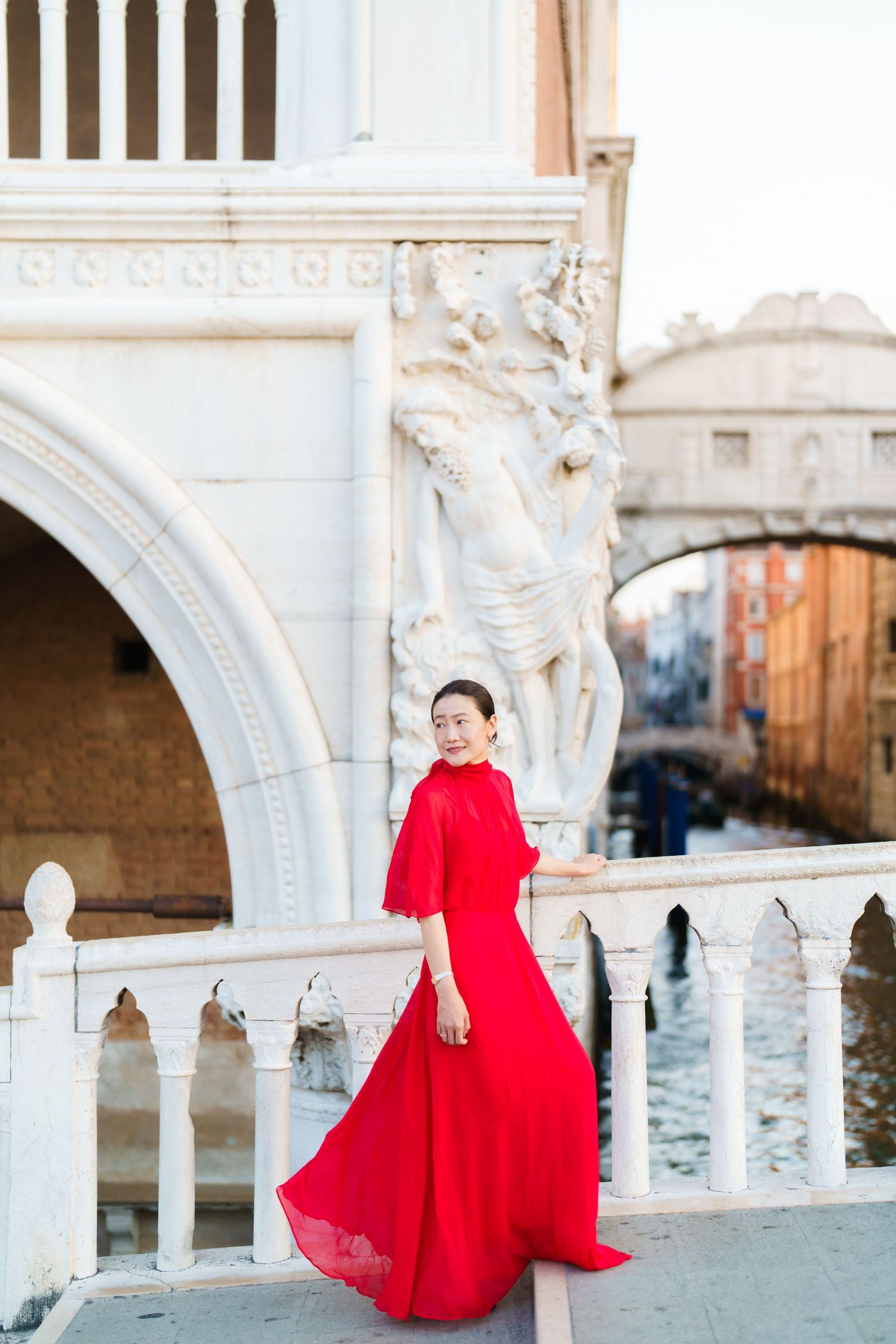 Woman in flowing red gown poses near ornate white marble balustrade with Venice canal and Bridge of Sighs behind her.