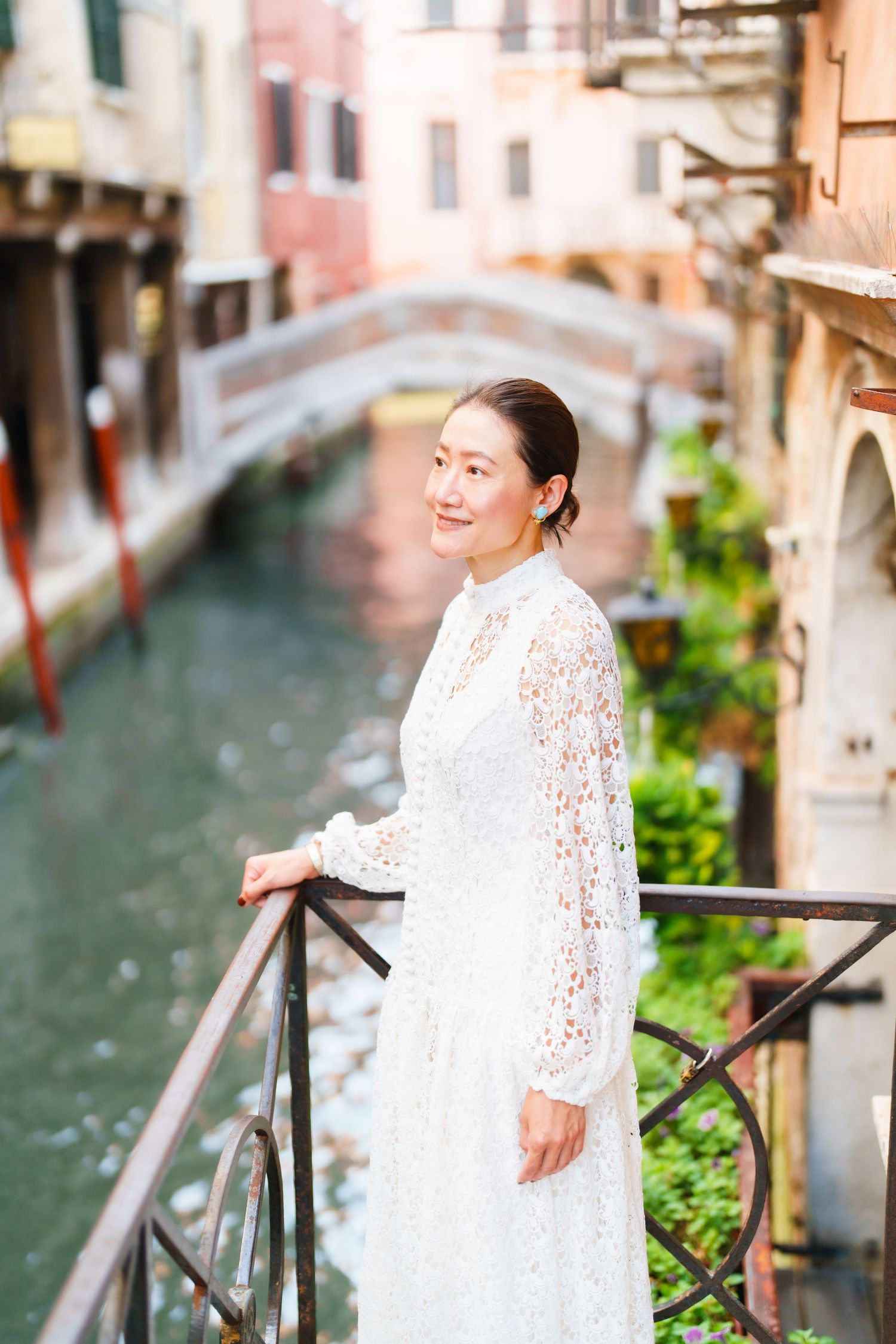 A woman in a white lace dress stands by a railing overlooking a scenic Venice canal with a bridge backdrop.