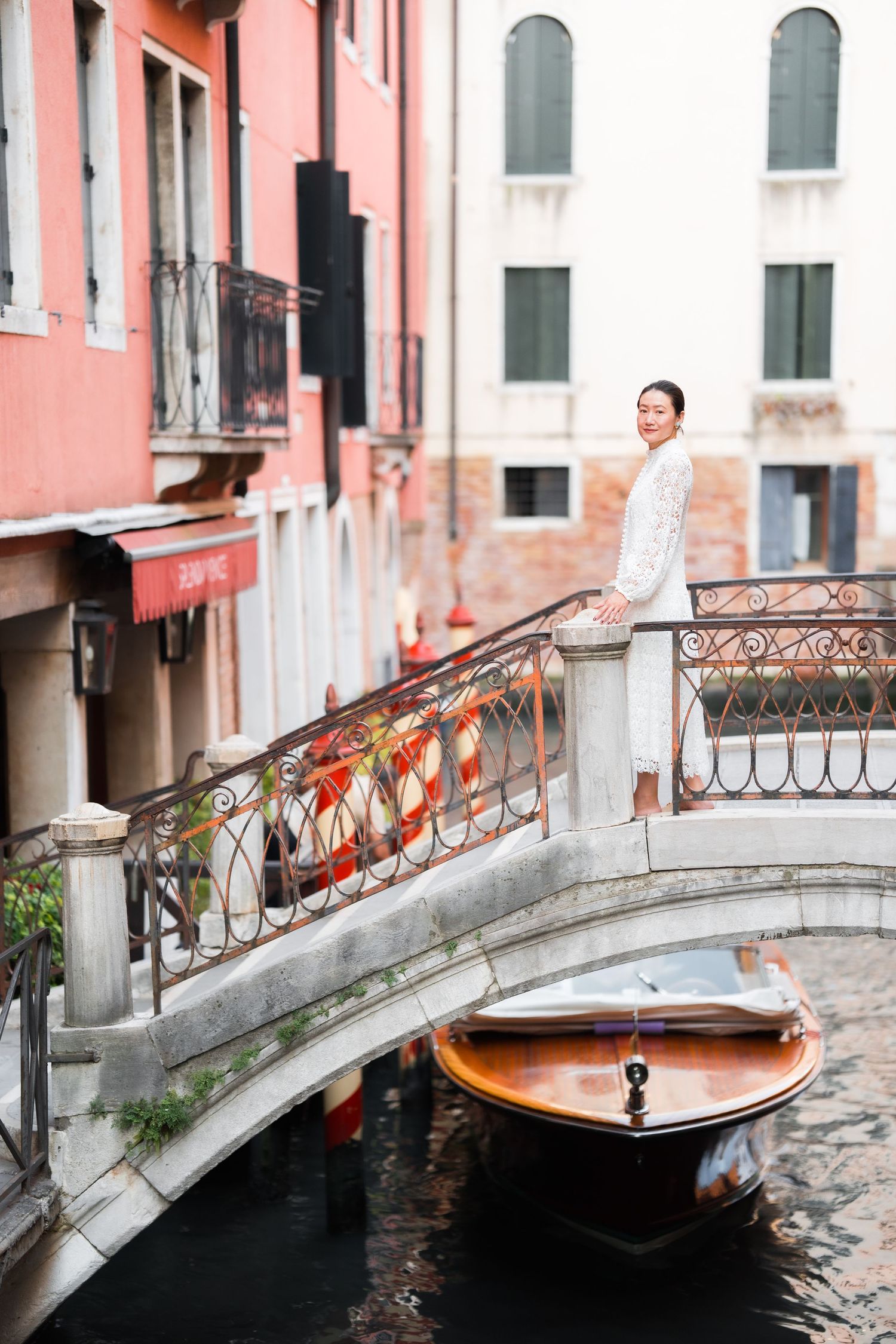 Woman in white dress standing on ornate bridge over Venice canal with wooden boat and colorful buildings.