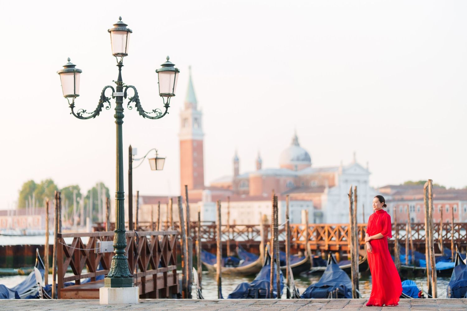 Woman in red dress stands by Venice waterfront with gondolas and San Giorgio Maggiore church in background.