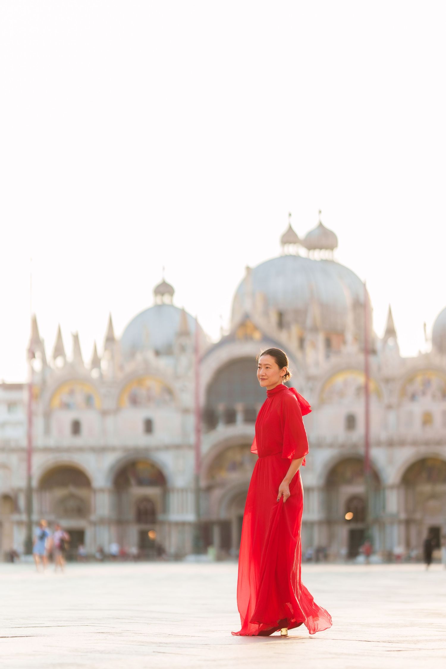 Woman in red gown poses in St. Mark's Square, Venice, with the iconic Basilica di San Marco in the background.