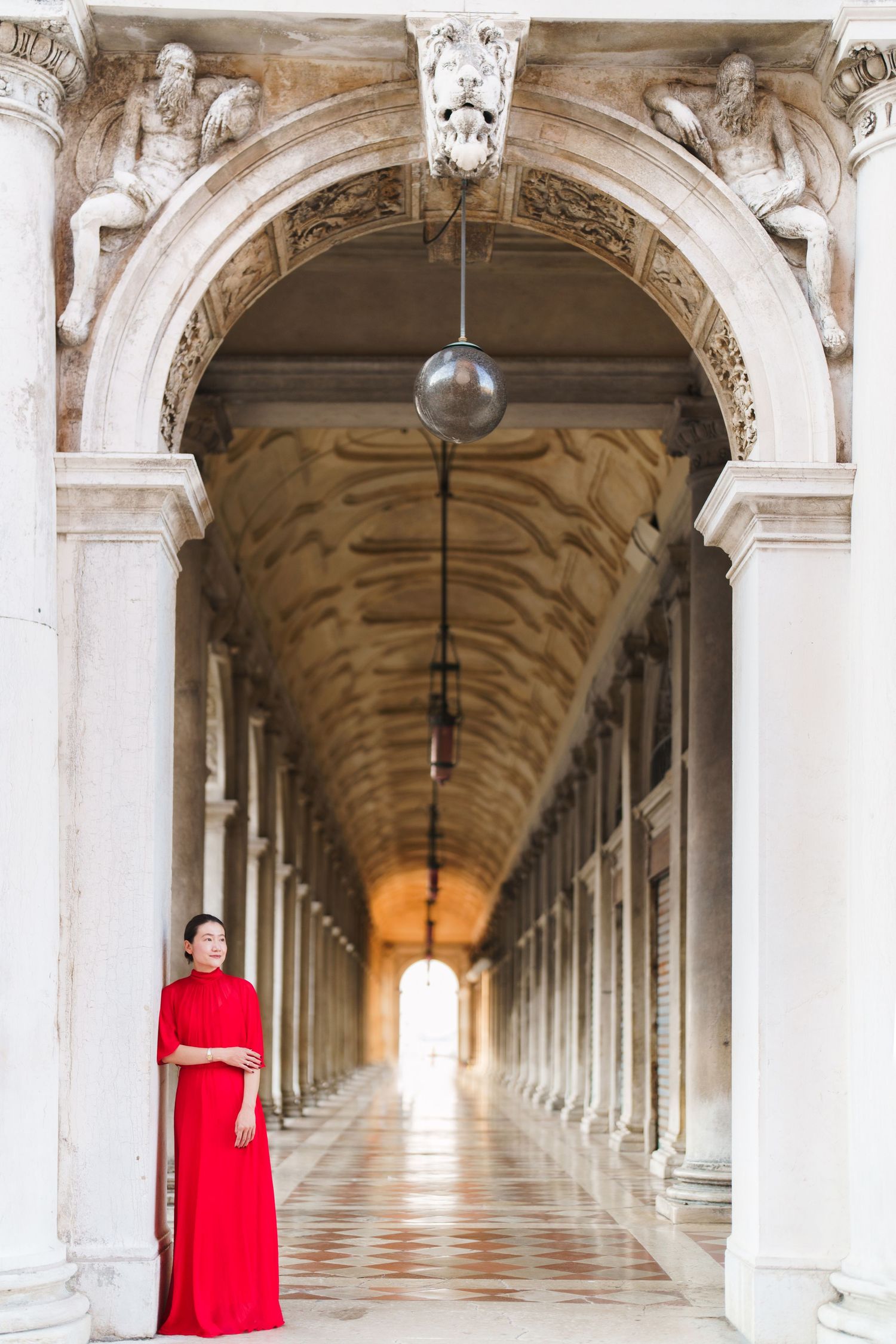 Woman in red dress standing in elegant white marble archway corridor with ornate columns and decorative ceiling.