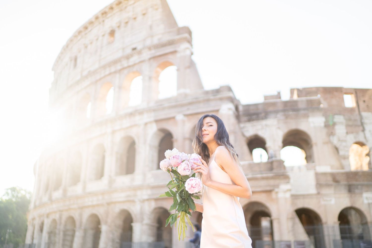 Woman in white dress holding pink roses in front of the Colosseum in Rome, Italy.