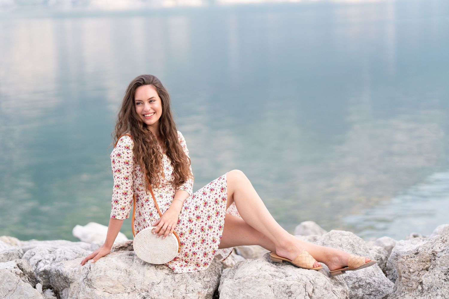 Smiling woman in floral dress sitting on rocky shore by calm turquoise water, holding a round white bag.