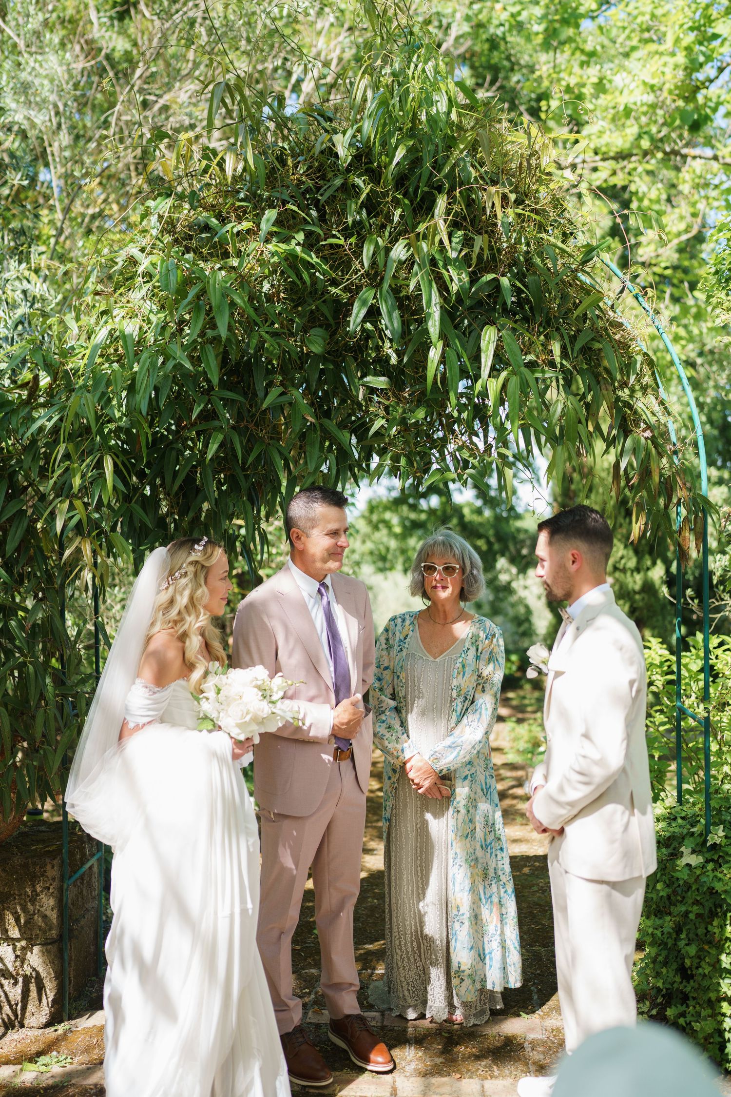 Bride and groom exchange vows under lush green arch with officiant in sunny outdoor garden wedding ceremony.