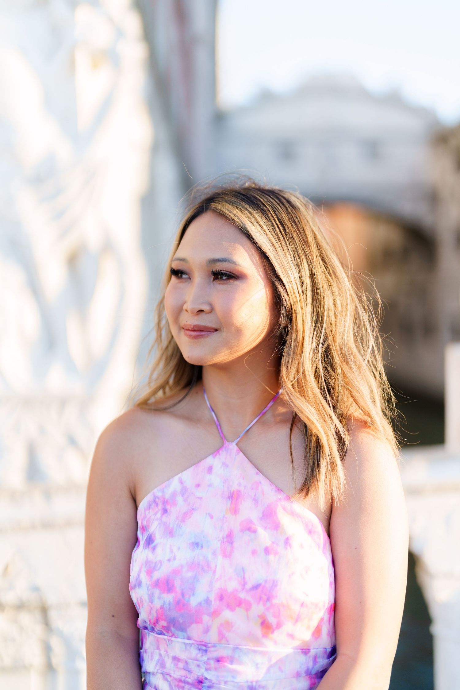 Young woman in pink floral halter dress poses outdoors near ornate white stone architecture in soft golden light.