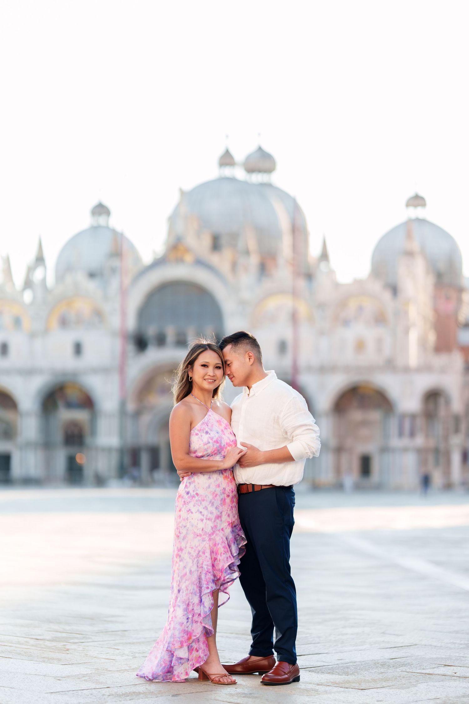 Couple embracing in St. Mark's Square, Venice, woman in pink floral dress, ornate Basilica di San Marco in background.
