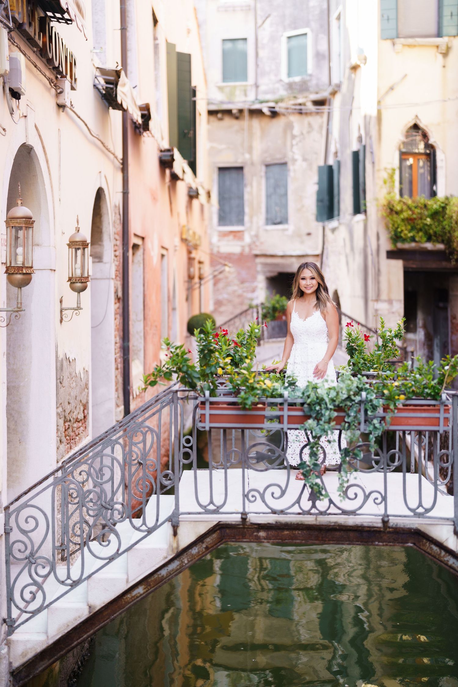 Bride in white dress standing on ornate bridge over Venice canal surrounded by colorful flowers and historic buildings.