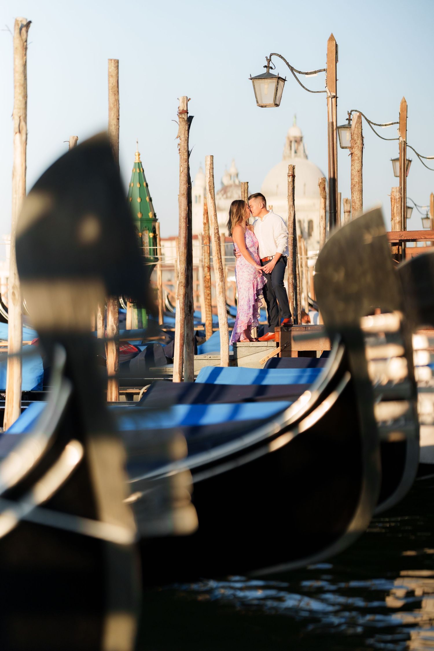 Romantic couple embracing on a Venice gondola dock with historic church and wooden mooring poles in background.