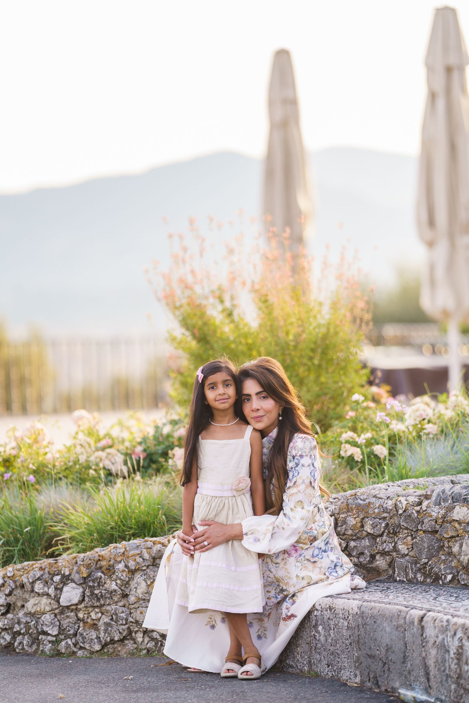 Woman and young girl in elegant dresses sitting on stone wall with garden and mountains in background.