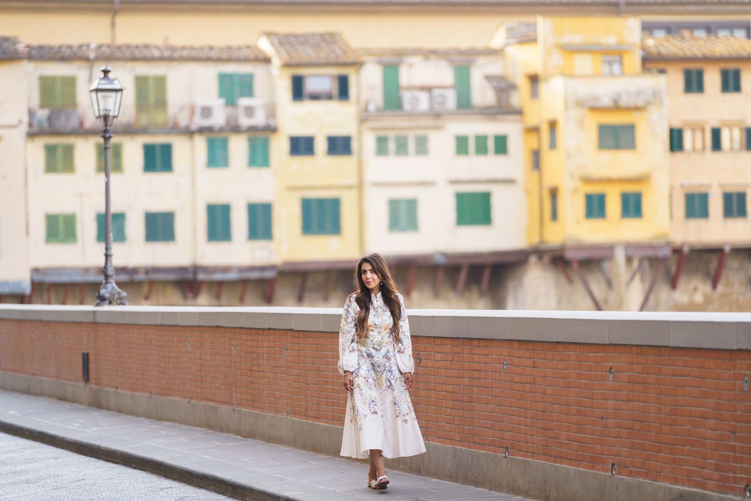 Woman in white floral dress walking on bridge with colorful Ponte Vecchio buildings in Florence, Italy.