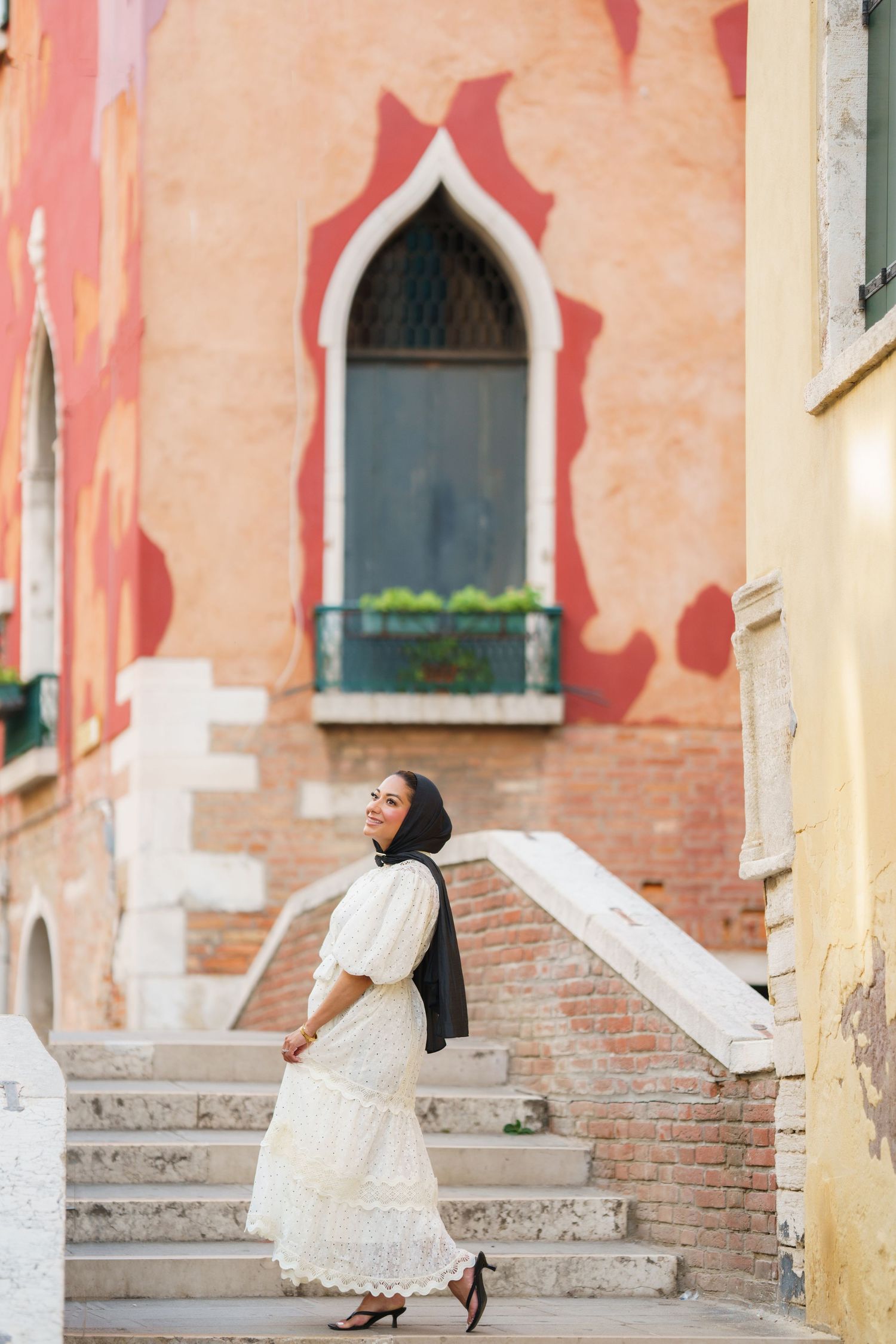 A woman in a white dress and black hijab walks up stone steps beside a colorful historic building in Venice, Italy.