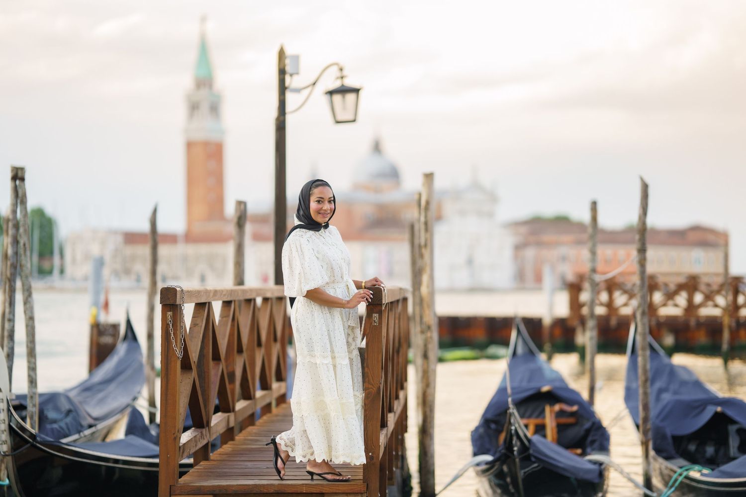Woman in white dress and hijab standing on wooden dock with gondolas and Venice skyline in background.