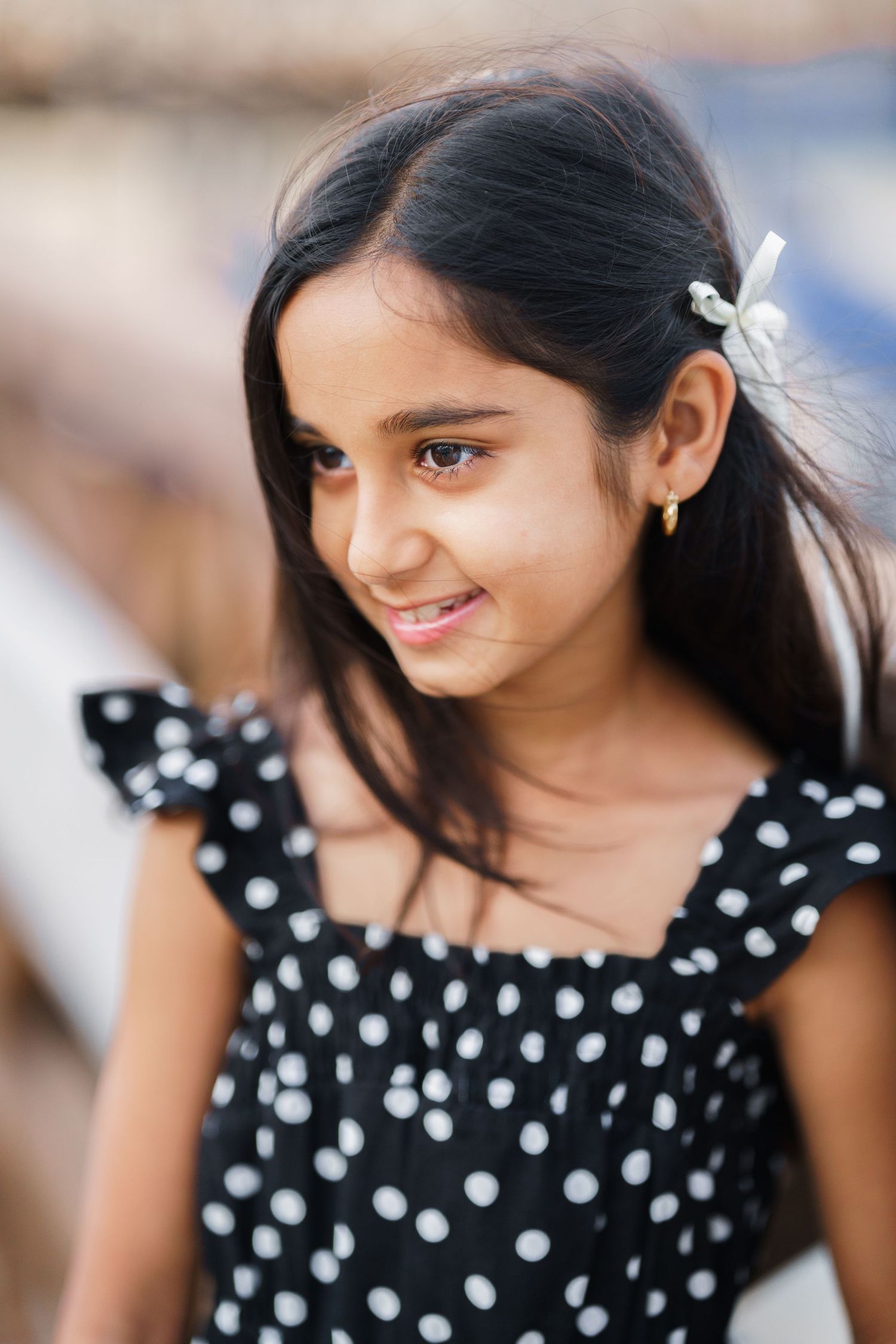Young girl in black and white polka dot dress with hair clip smiling softly outdoors.