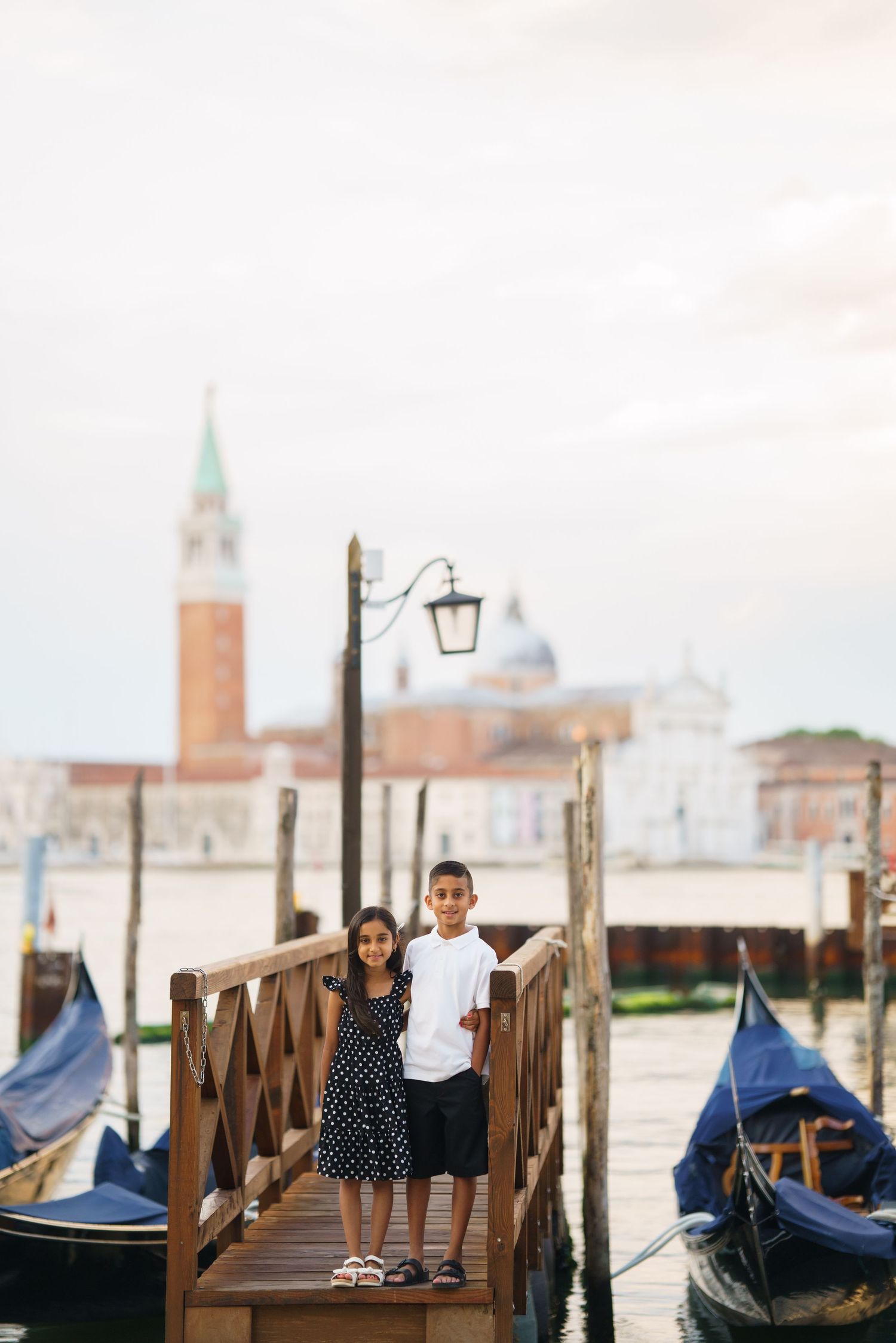 Two children pose on a wooden dock in Venice, Italy, with gondolas and San Giorgio Maggiore church in the background.