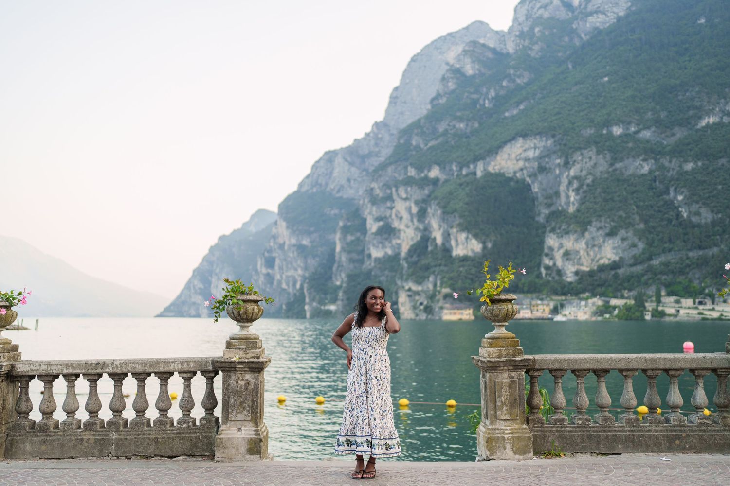 Woman in summer floral dress standing on lakeside terrace with Lake Garda mountains in background.