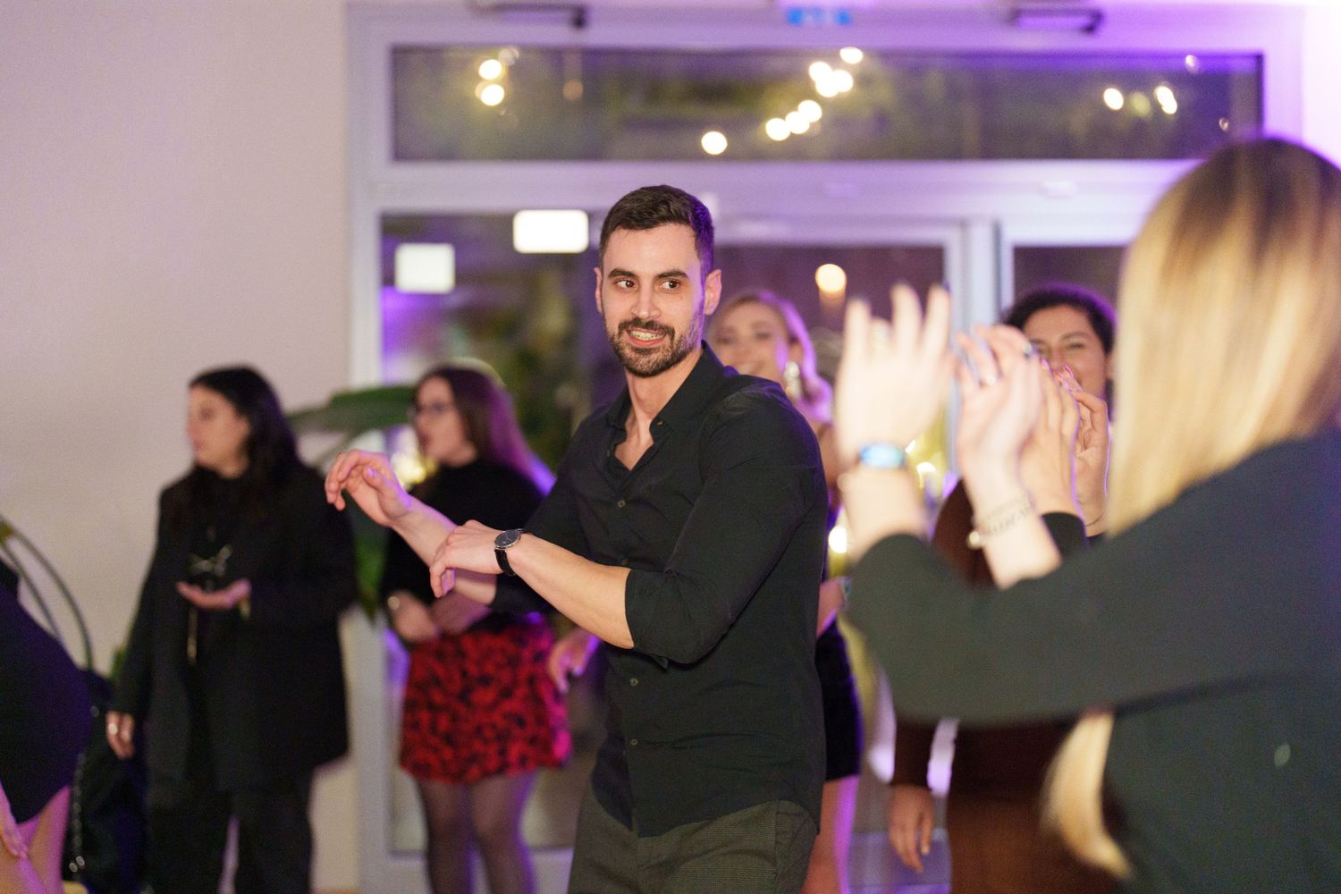 Man in black shirt dancing at a lively indoor party with guests clapping and celebrating in the background.
