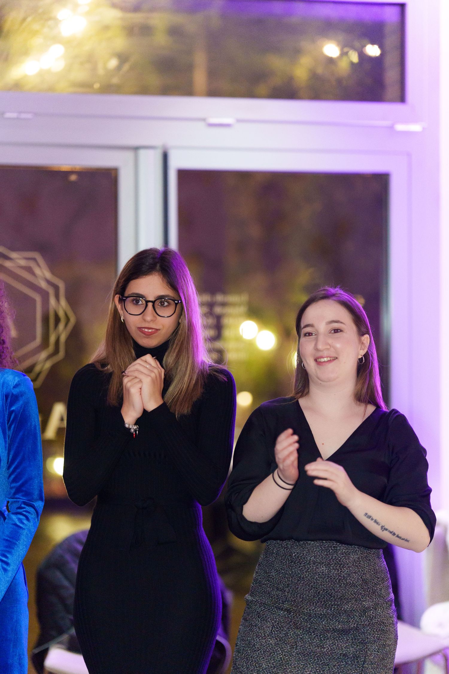 Two women in black outfits clapping and smiling at an indoor event with purple lighting.