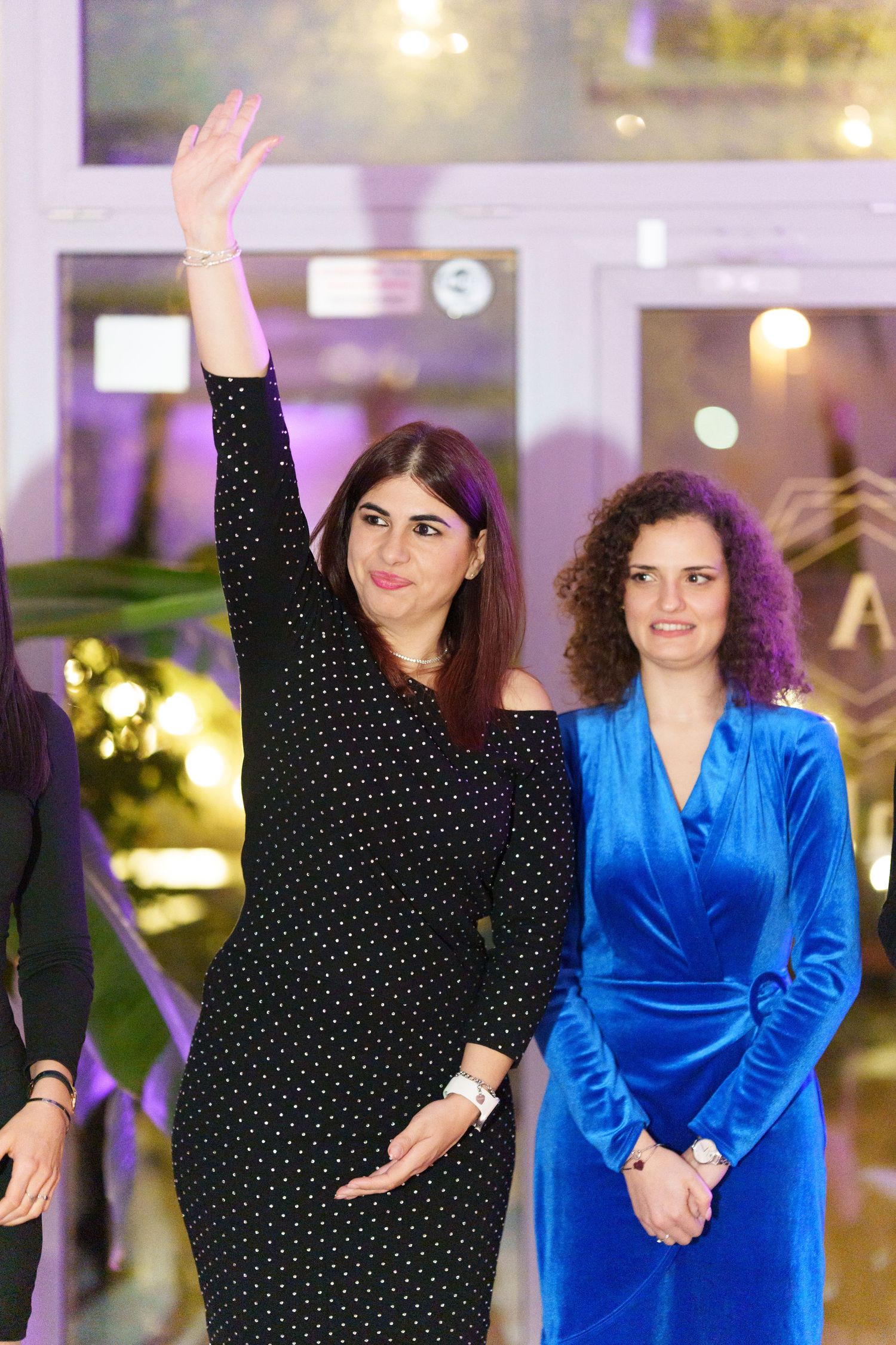 Two women at a formal event, one in black polka dot dress waving, another in blue velvet wrap dress smiling.