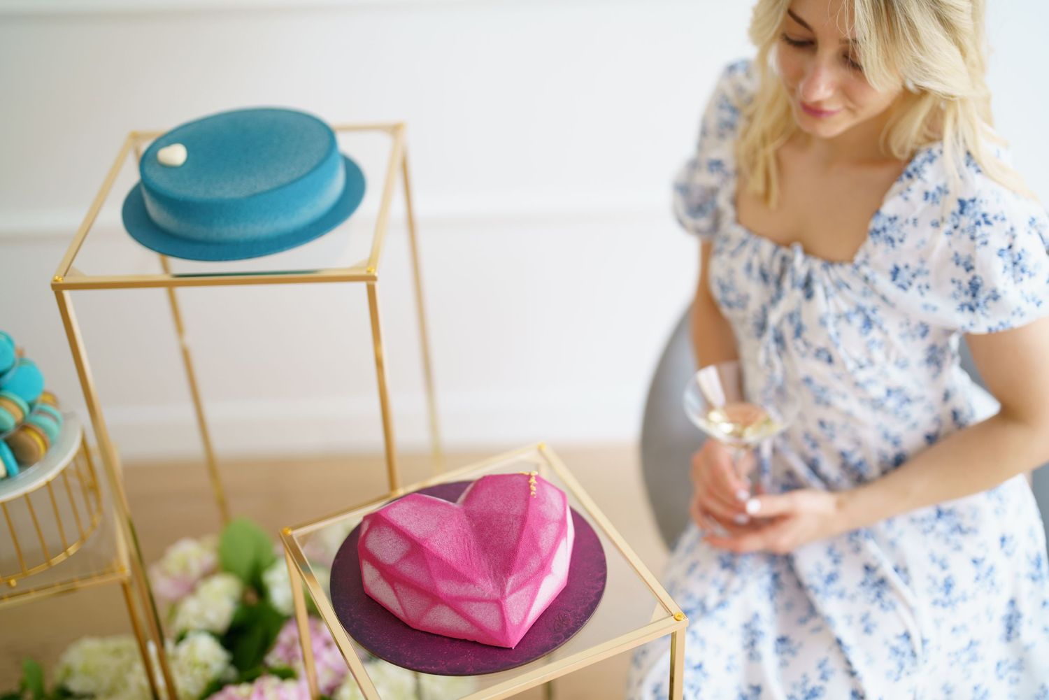 A blonde woman in a floral dress admires colorful heart-shaped cakes on gold stands at an elegant dessert display.