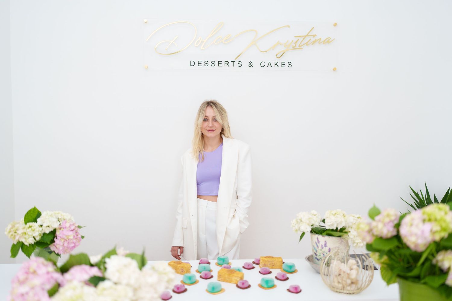 Woman in white blazer stands before a desserts and cakes shop display with colorful macarons and floral arrangements.