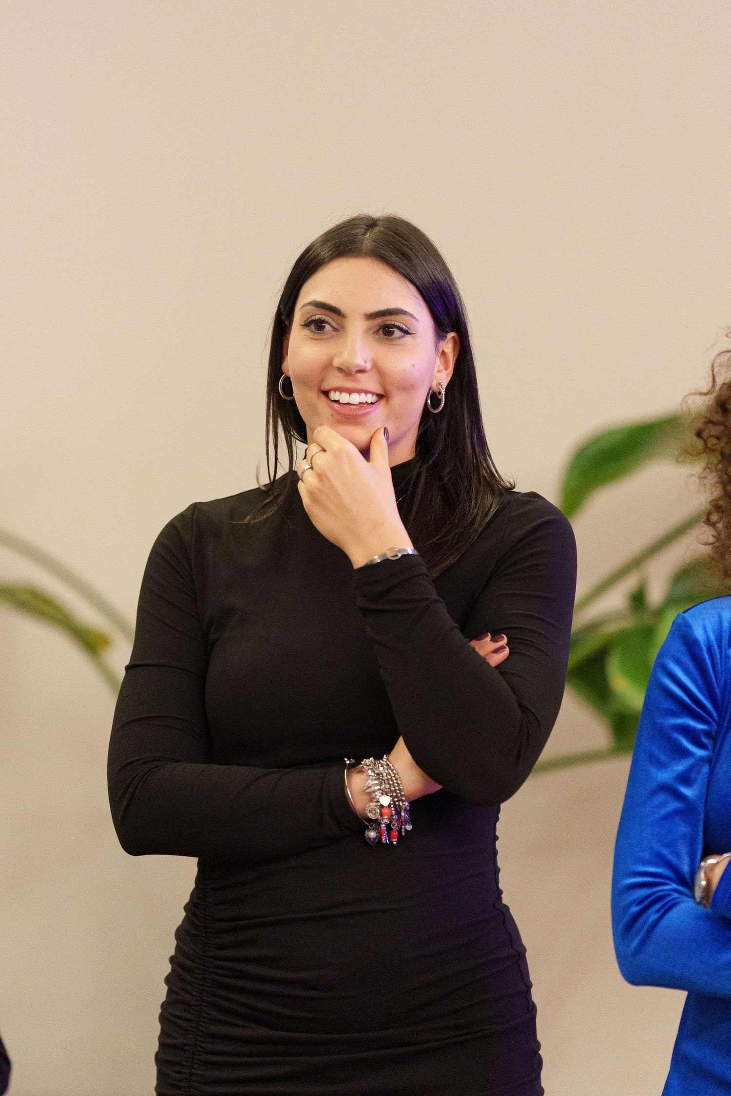 Young woman in black dress smiling thoughtfully with hand on chin, wearing bracelets, standing indoors.