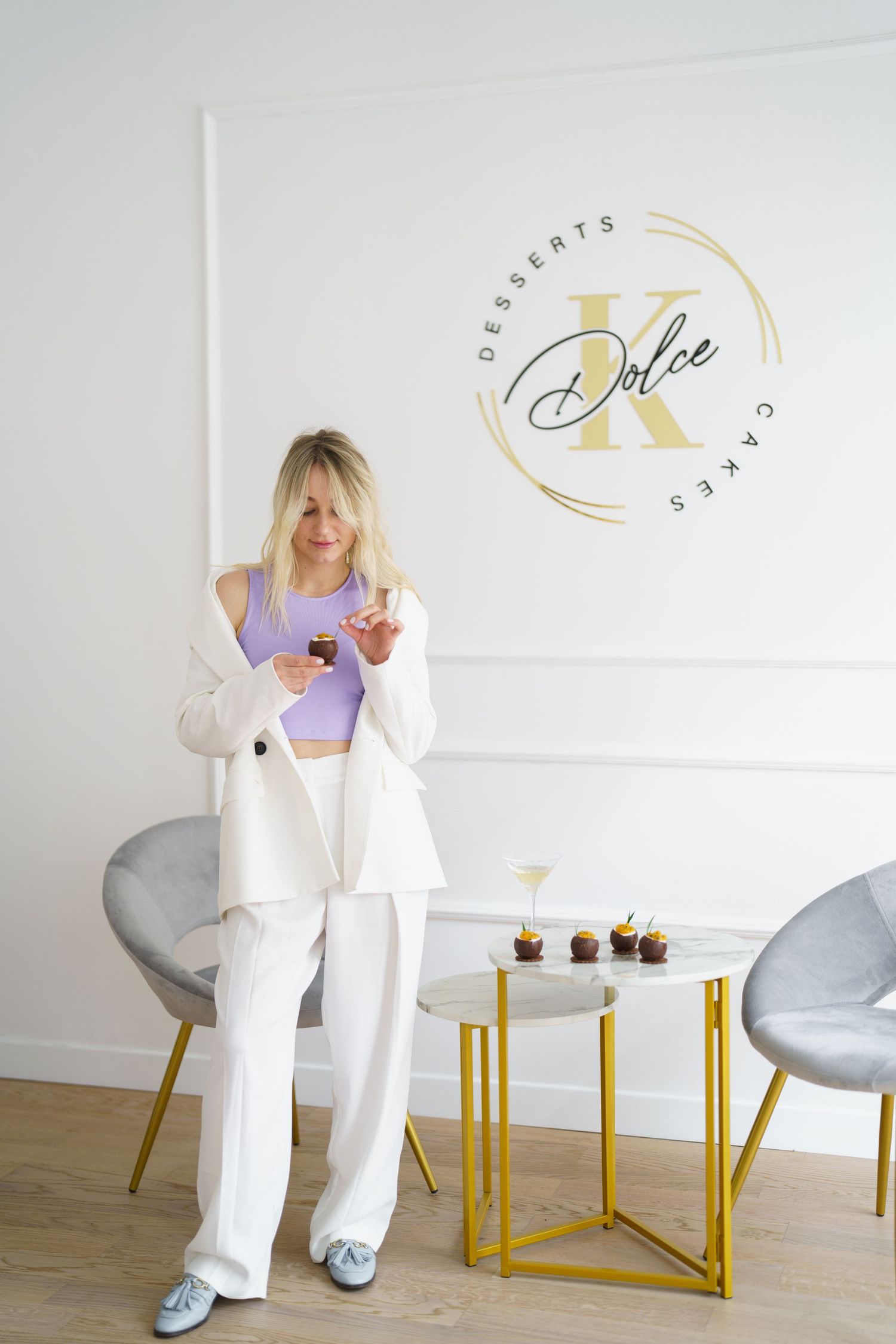Blonde woman in white suit holding cupcake beside gold table in Dolce K Desserts and Cakes bakery showroom.