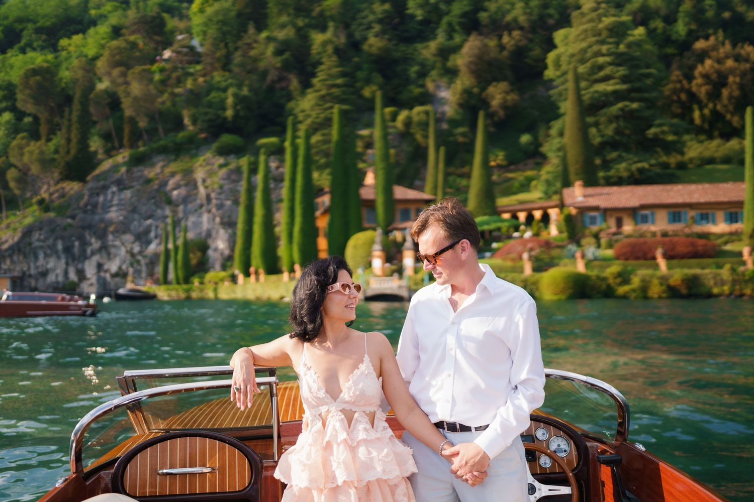 Couple in elegant attire sitting on a wooden boat on Lake Como, Italy, with lush green hills and villa in background.