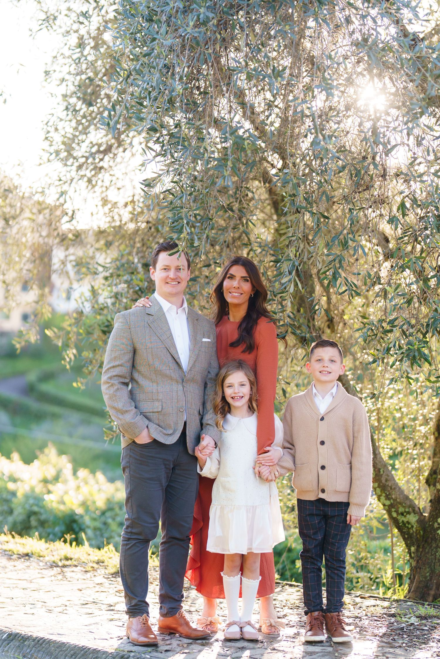 Happy family of four posing outdoors under an olive tree in elegant coordinated outfits on a sunny day.