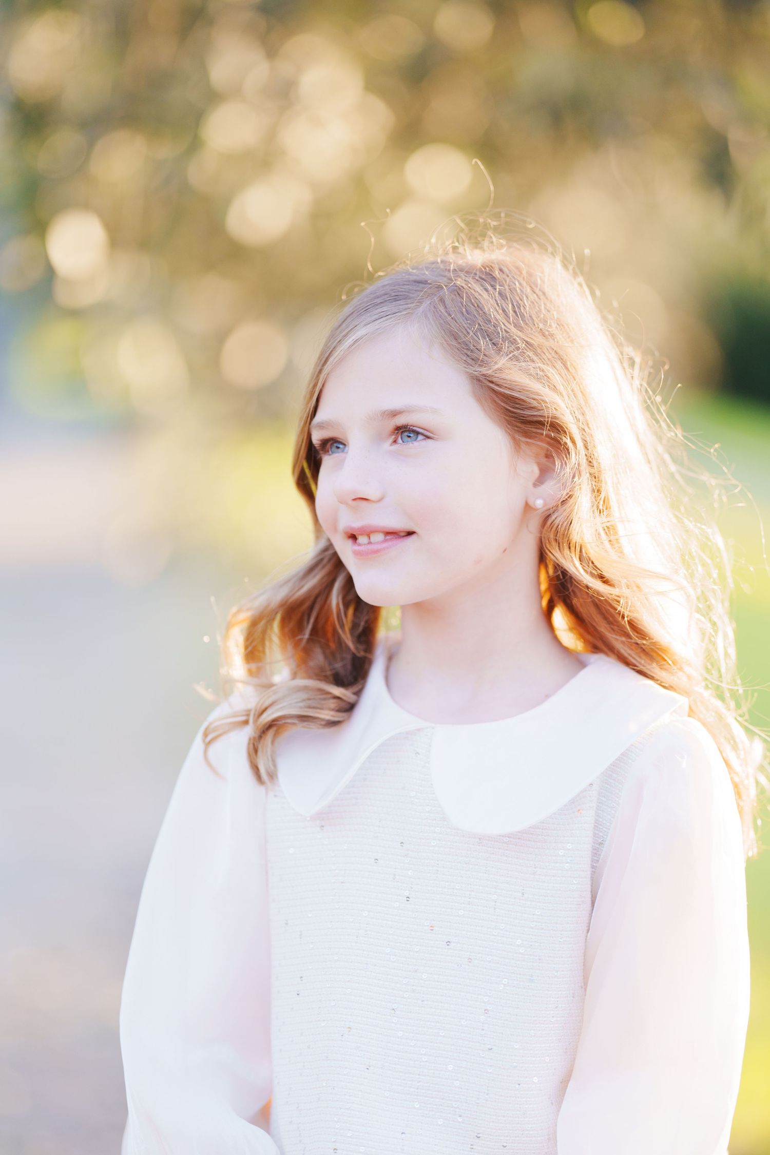 Young girl with long auburn hair and blue eyes wearing a white dress, photographed outdoors in soft golden light.
