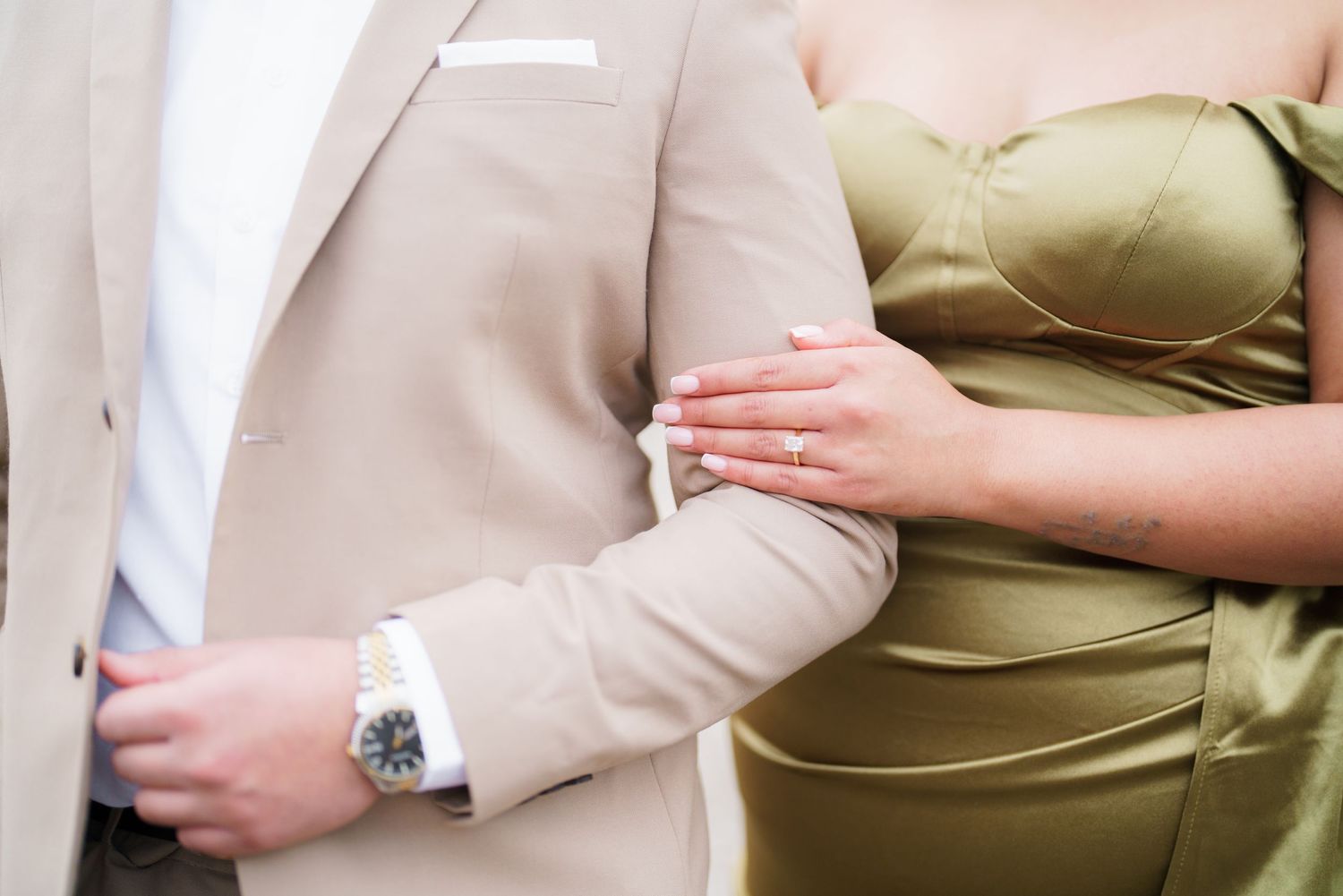 Couple in elegant attire, man in beige suit with watch, woman in olive dress, showing engagement ring.