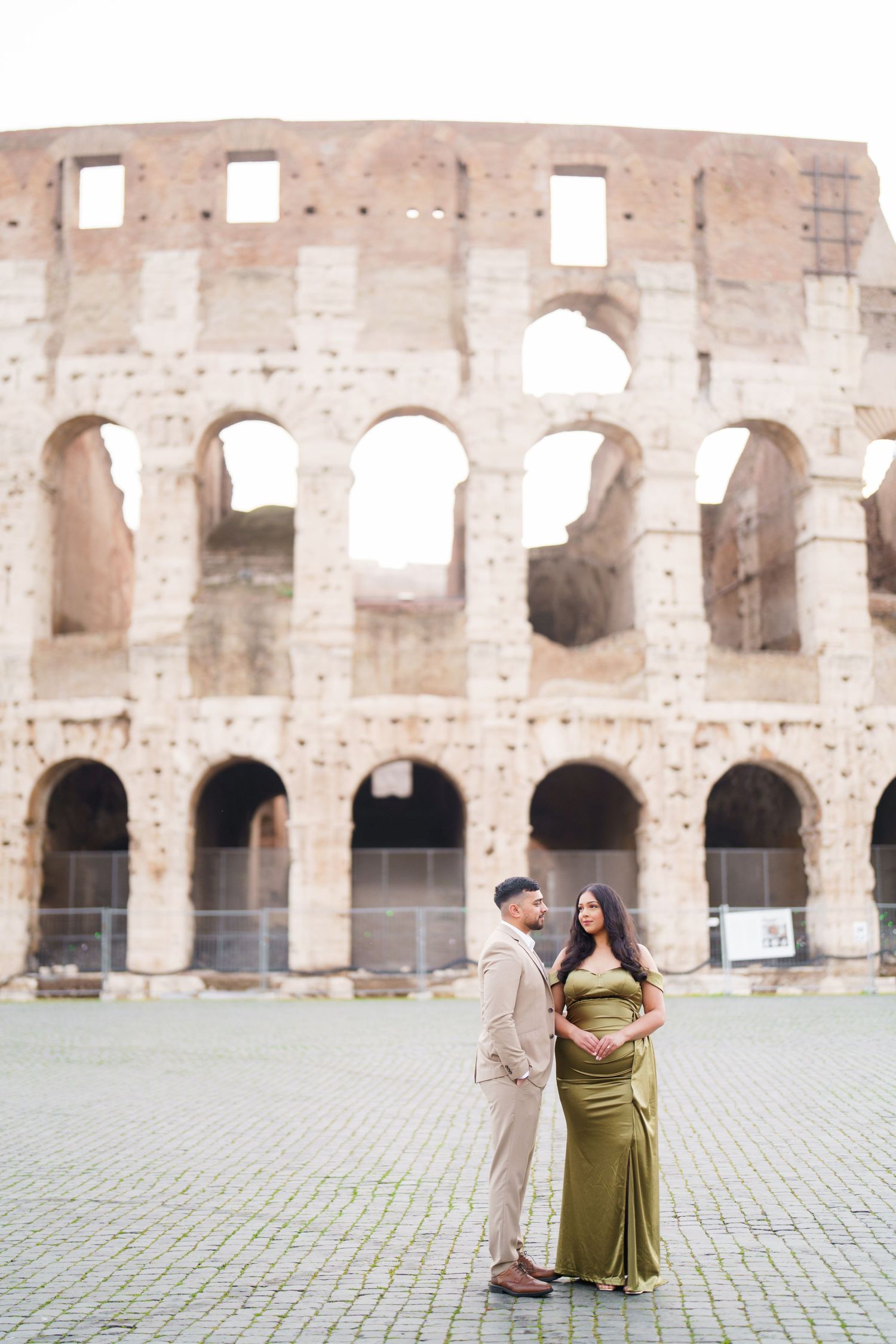 Couple poses before the Colosseum in Rome, woman in green gown cradling baby bump, man in beige suit.
