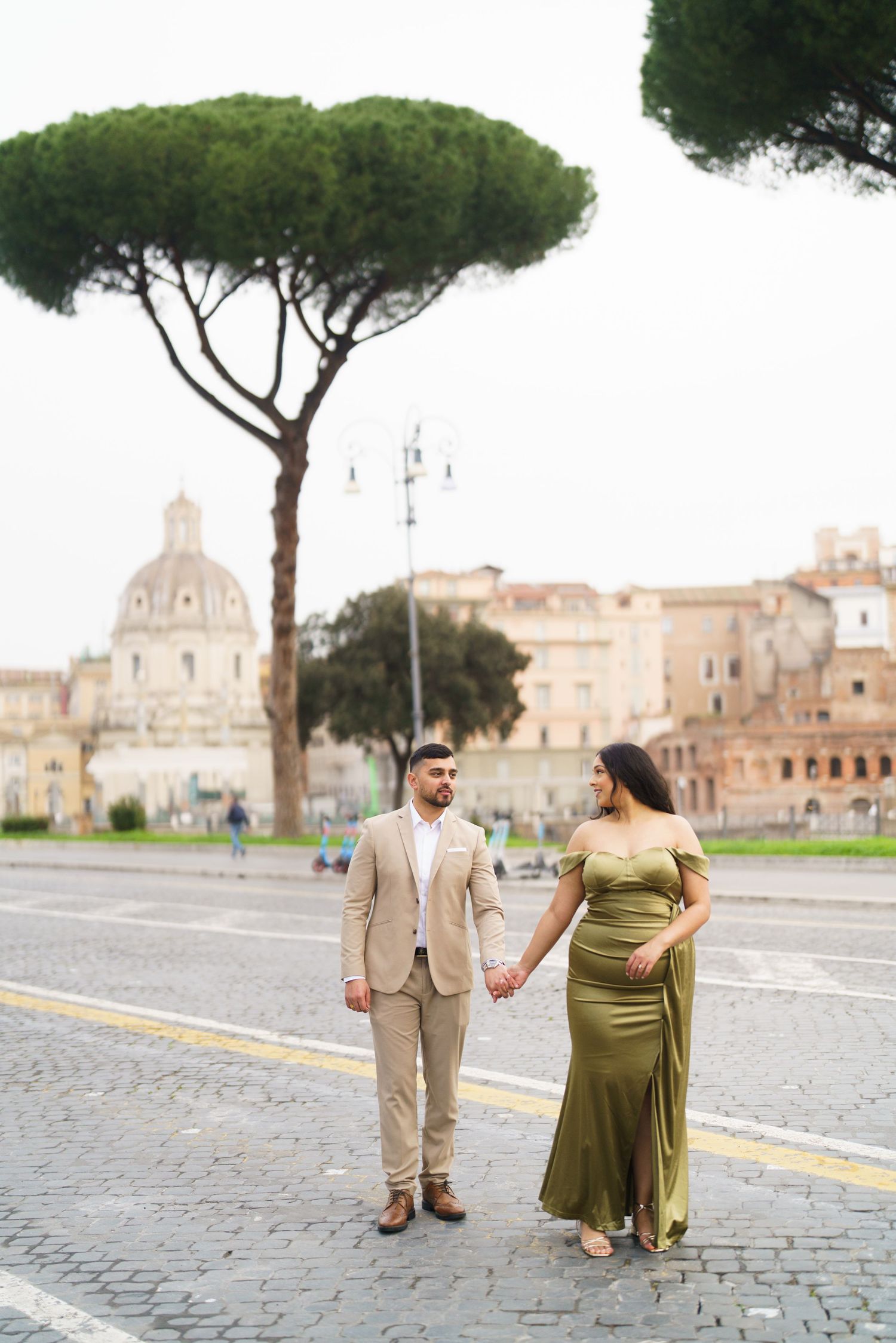 Couple holding hands in Rome, woman in green gown and man in tan suit, with historic ruins and pine trees behind them.